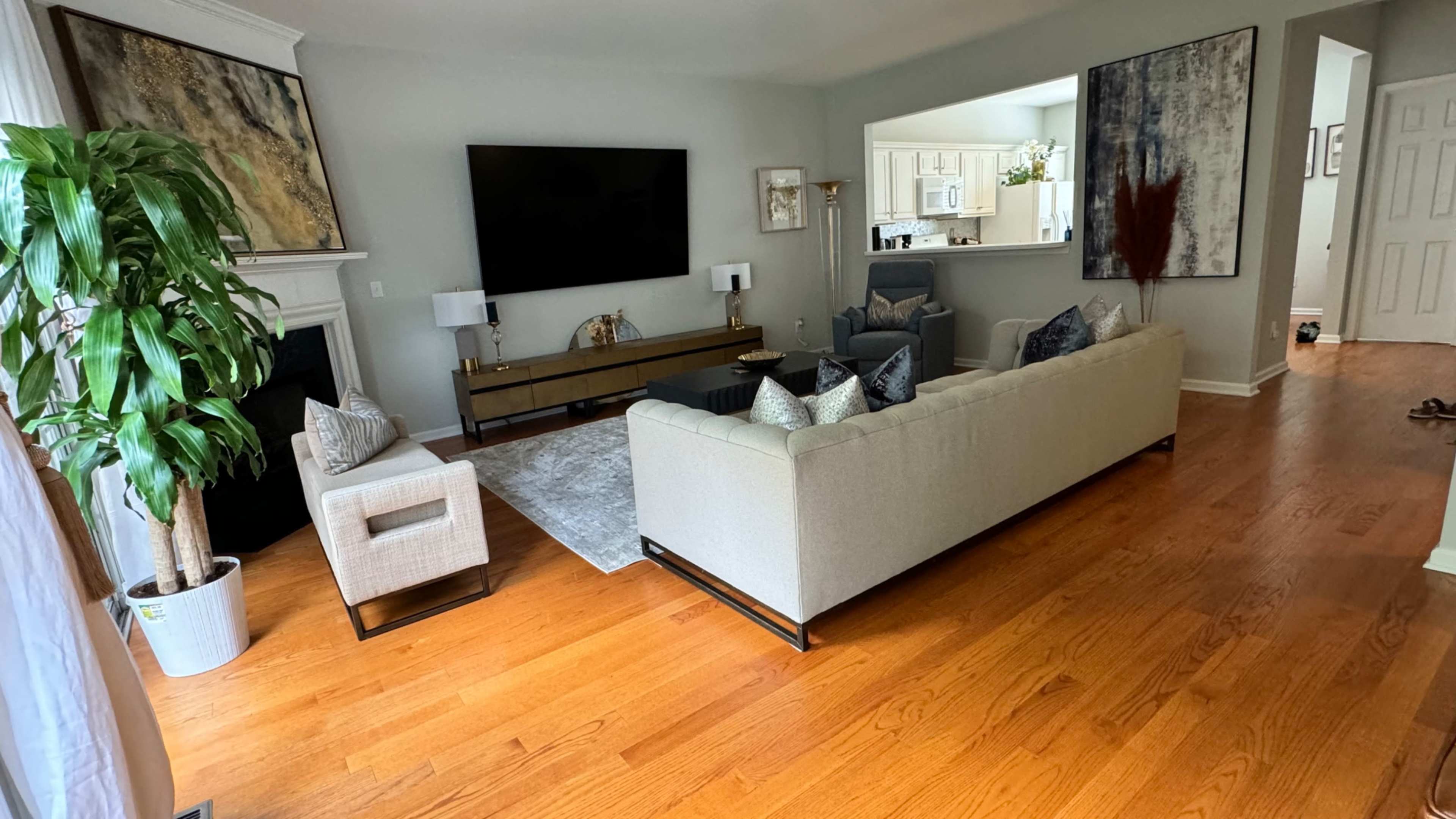 The living room features a large gray sofa, two armchairs, a wall-mounted television, and a dining area visible in the background.