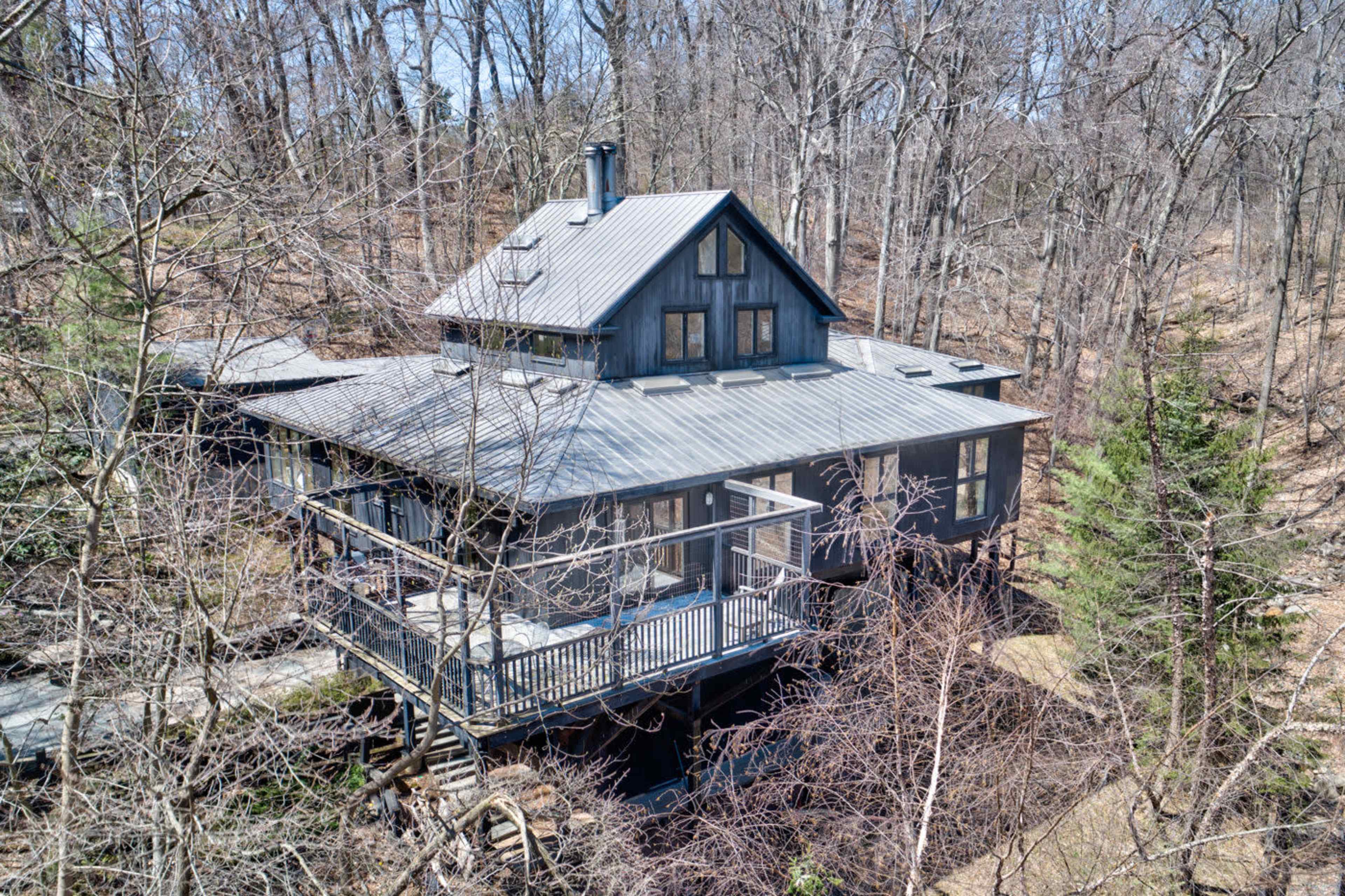 A modern black house with a sloped roof is situated in a wooded area, surrounded by bare trees and a deck extending from the front.
