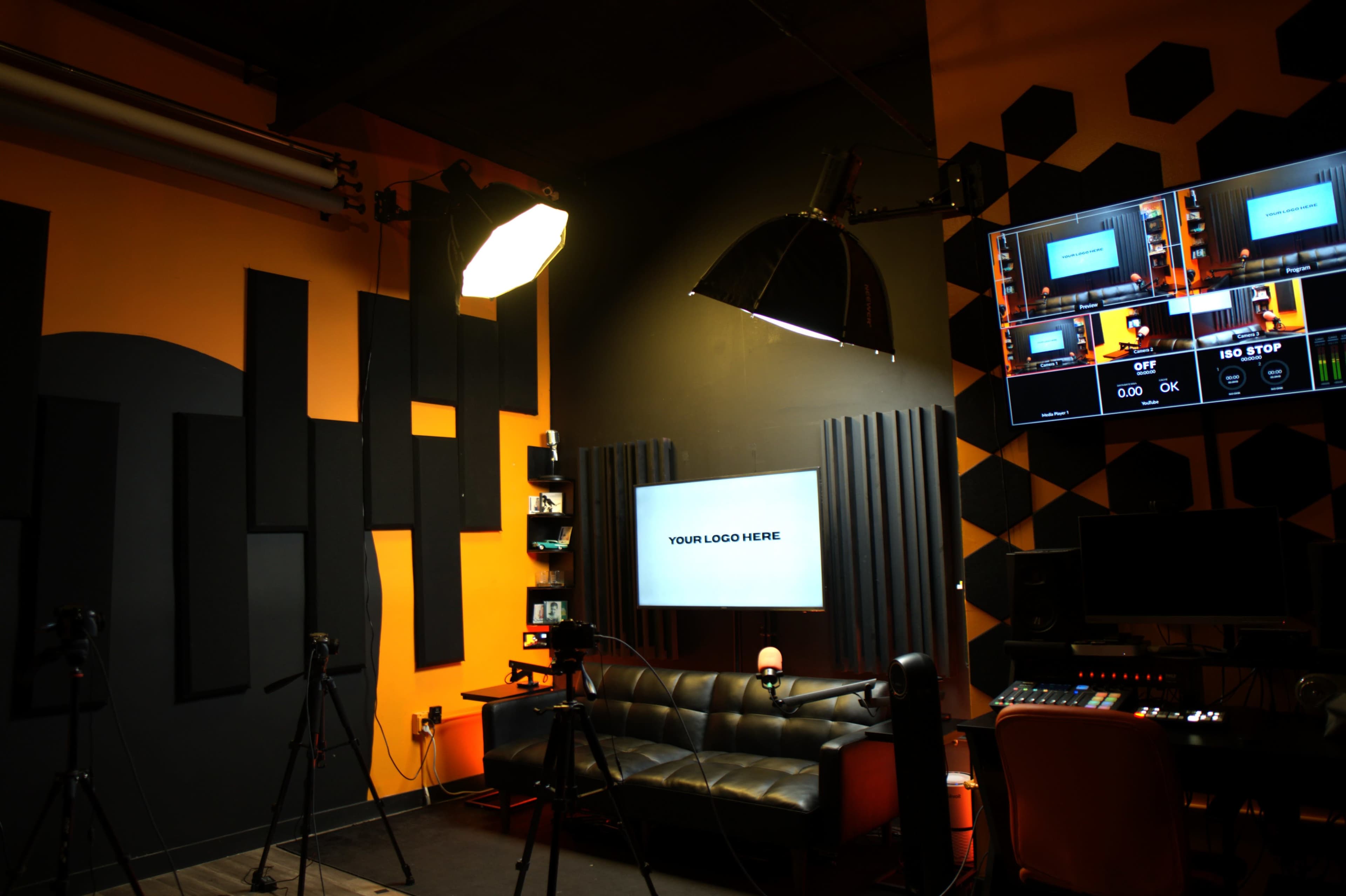 The image shows a modern studio setup with a black couch, screens displaying video feed controls, and orange and black geometric wall designs.