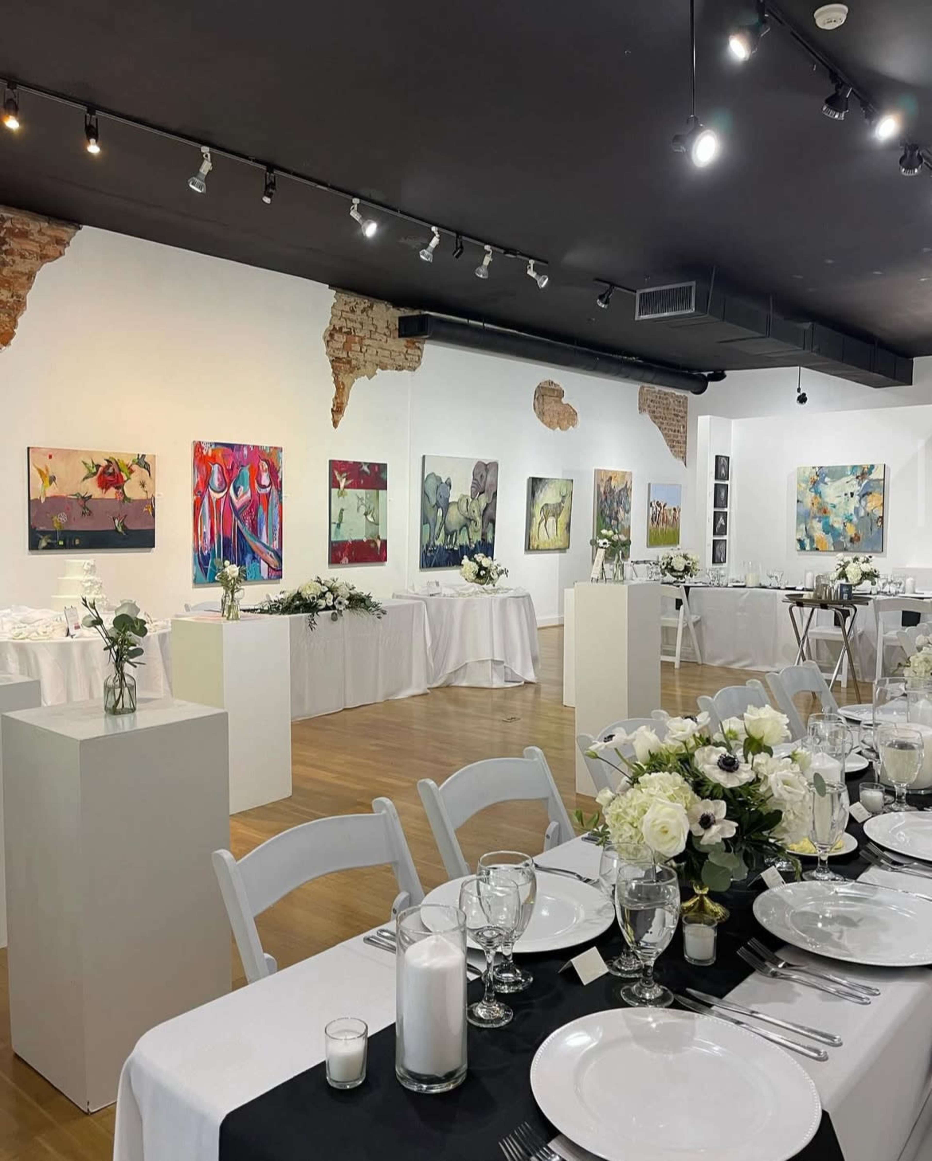 The image shows an art gallery setup for an event, featuring white tables adorned with floral centerpieces and various abstract paintings displayed on the walls.