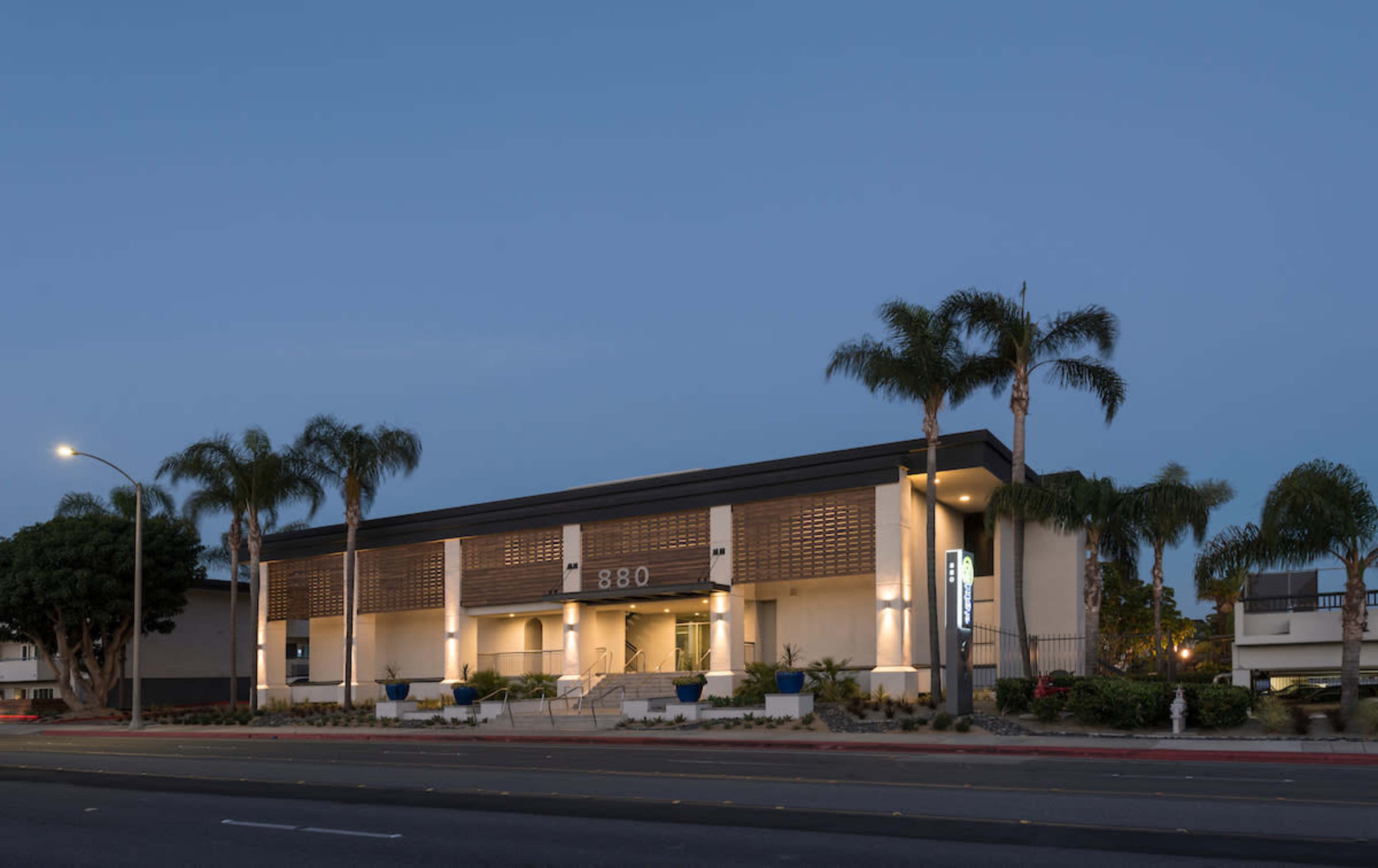 A modern, two-story building with a distinctive facade and palm trees in front, located on a city street at dusk.