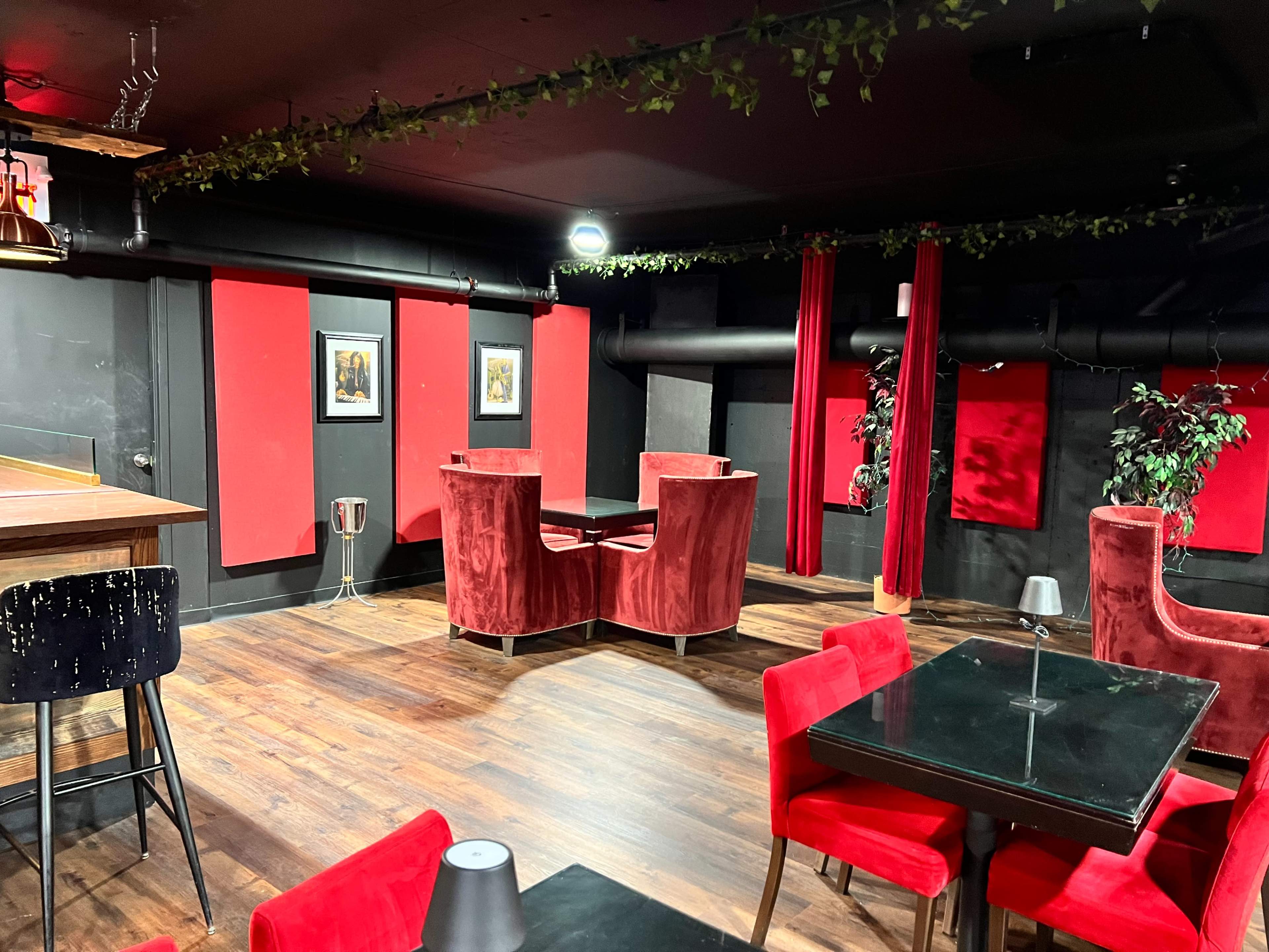 The image shows a dimly-lit lounge with red velvet chairs, black tables, and framed artwork on the red walls.