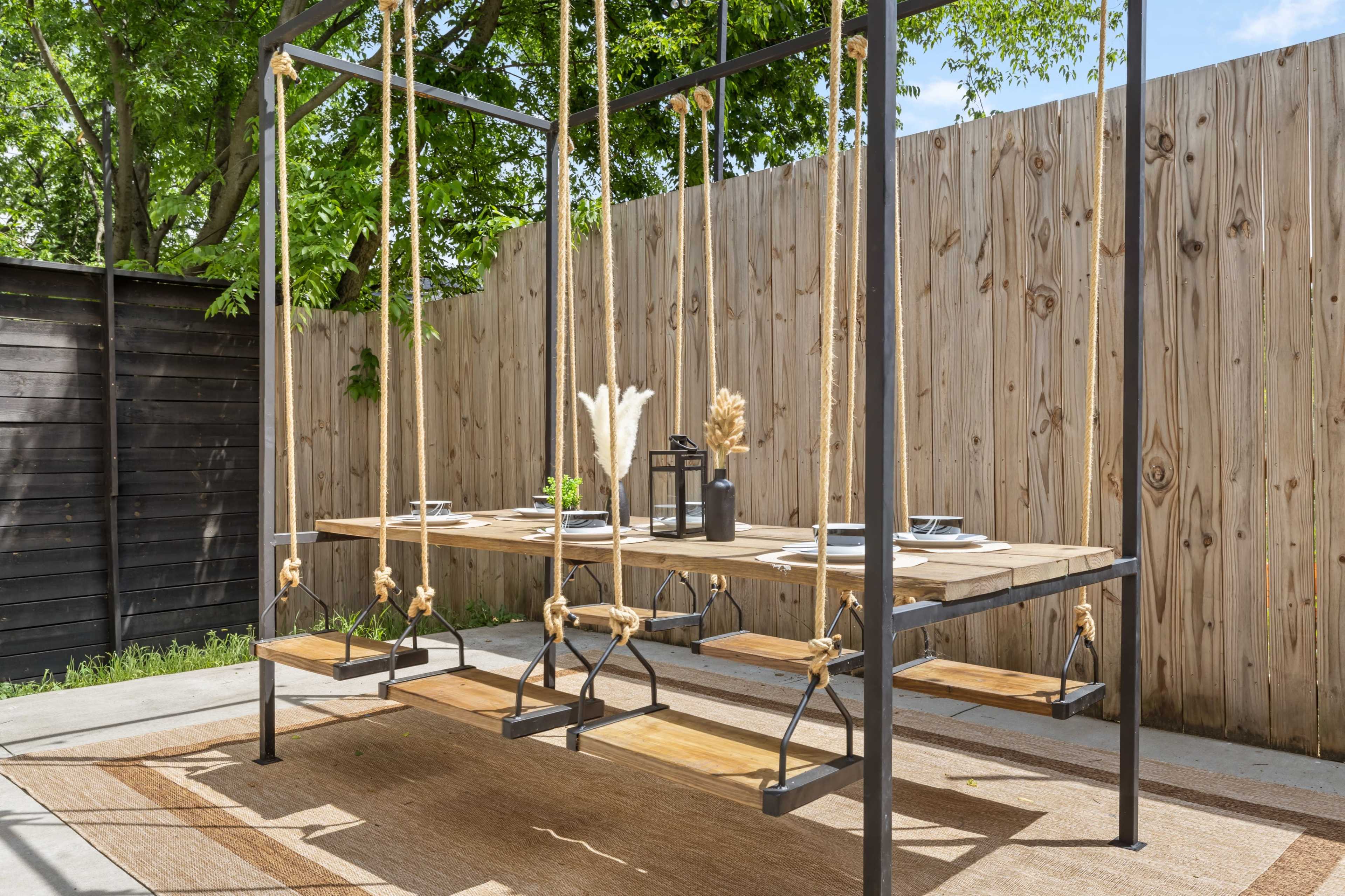 The image shows a wooden swing dining table supported by ropes under a shaded outdoor area, surrounded by a wooden fence.