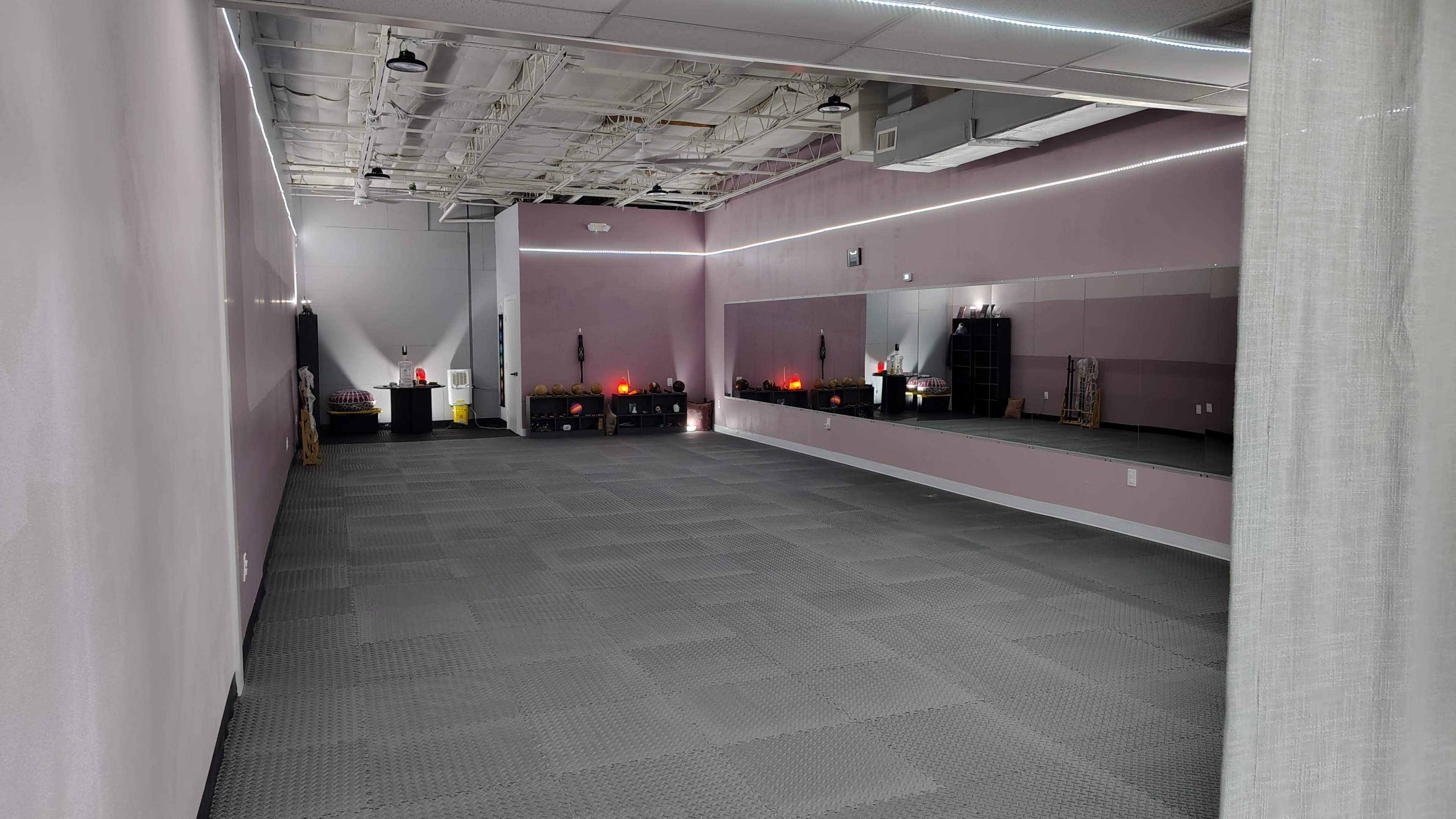 The image shows an empty, well-lit room with gray rubber flooring, soft pink walls, and mirrors along one side.
