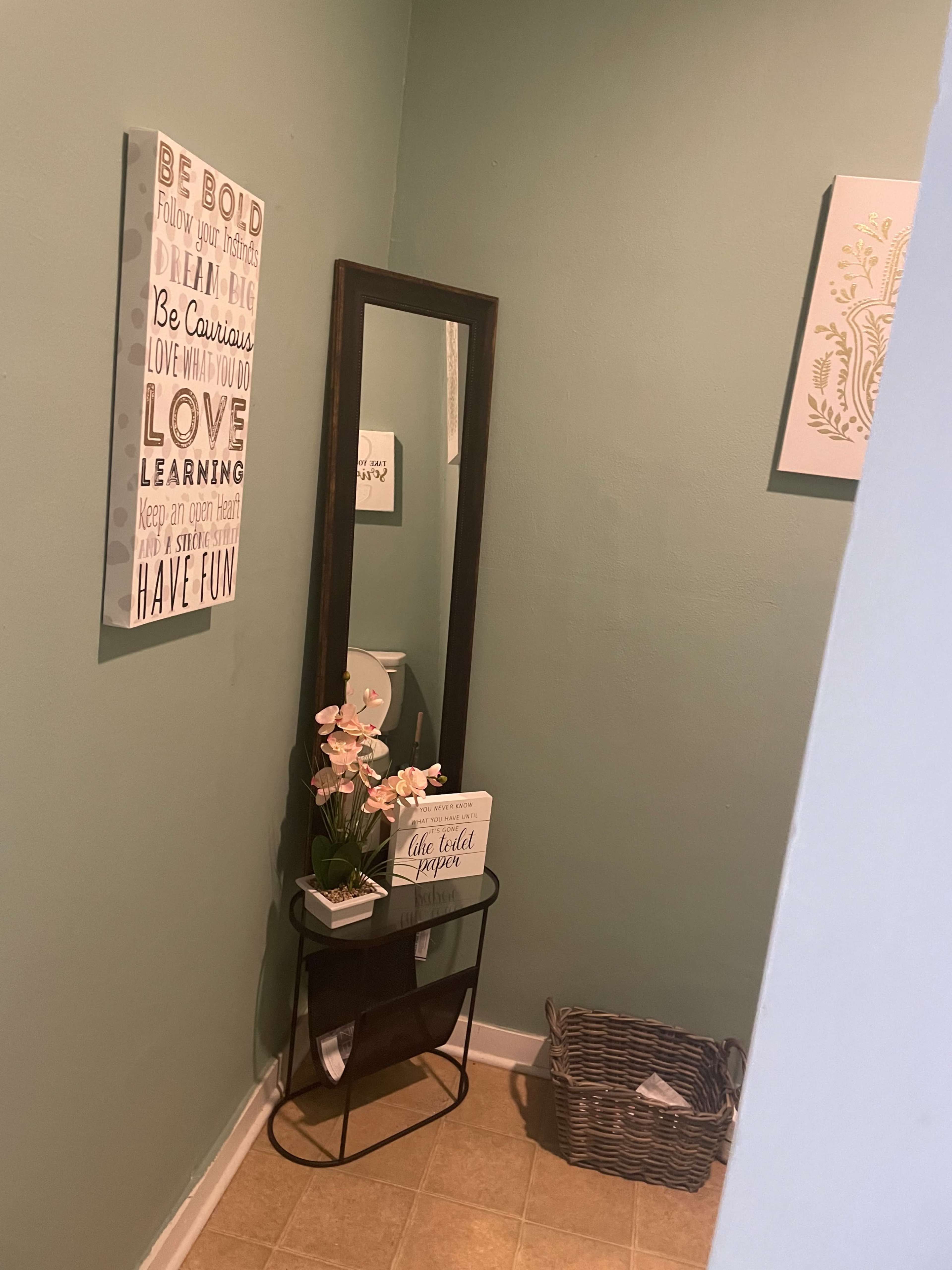 A corner of a room featuring a green wall, a large mirror, a decorative sign with motivational phrases, a small plant arrangement, and a wicker basket.
