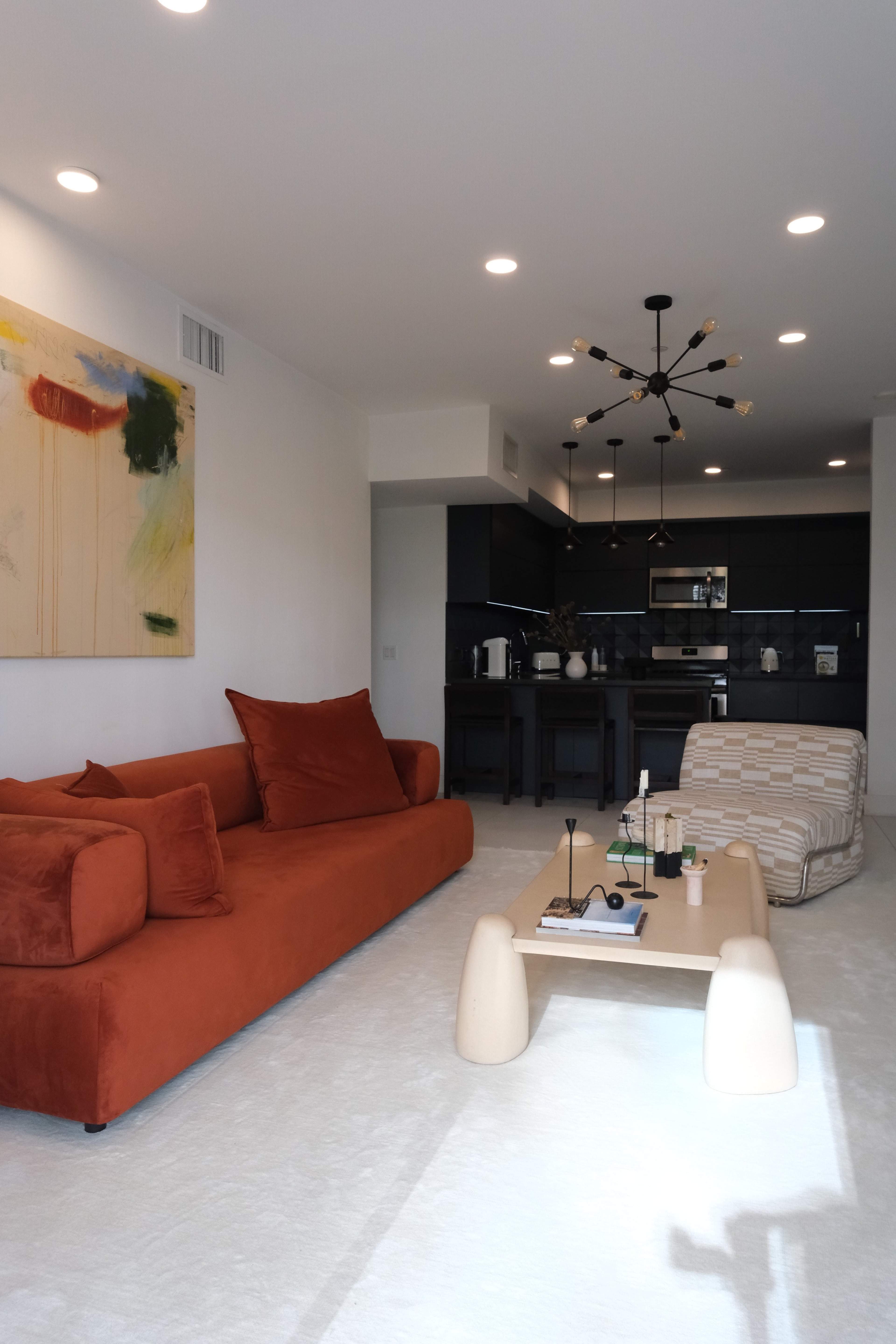 The image shows a modern living room featuring an orange sofa, a light-colored coffee table, and a kitchen area in the background with dark cabinetry.