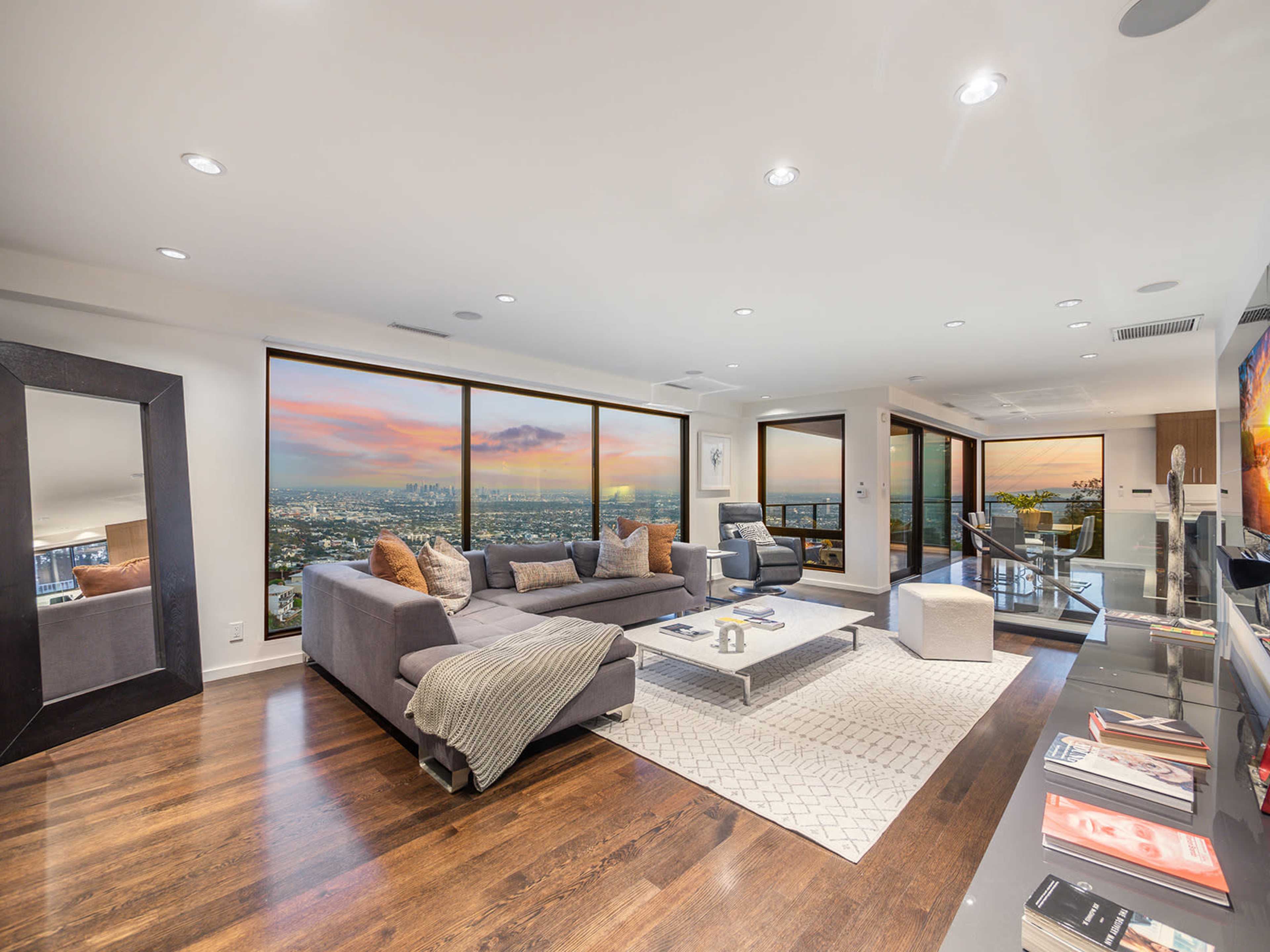 A modern living room features large windows with a view of the city skyline and sunset, furnished with a sectional sofa, coffee table, and a sleek entertainment unit.