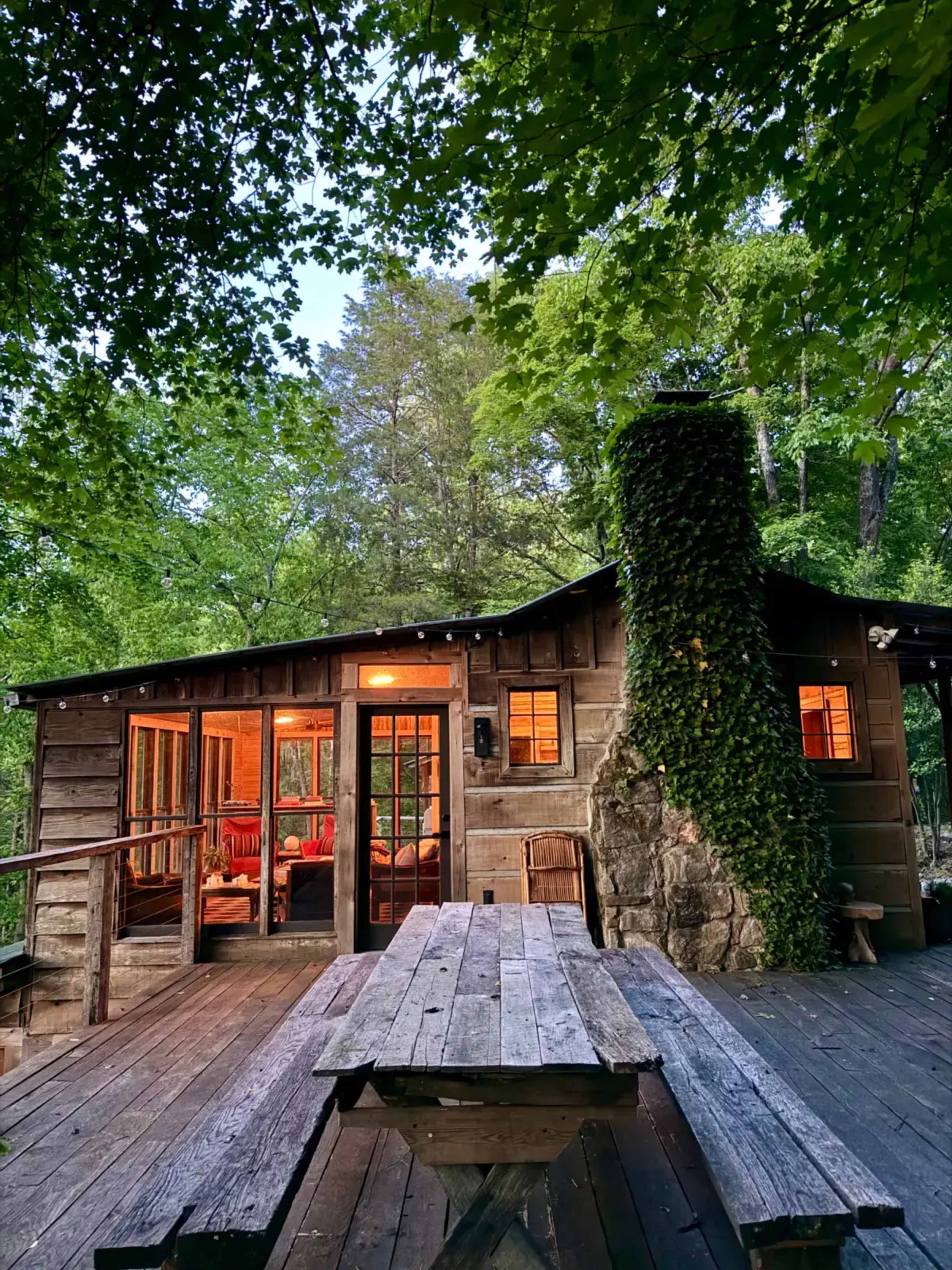 A wooden cabin with a stone chimney sits among lush green trees, featuring large windows that illuminate the interior.