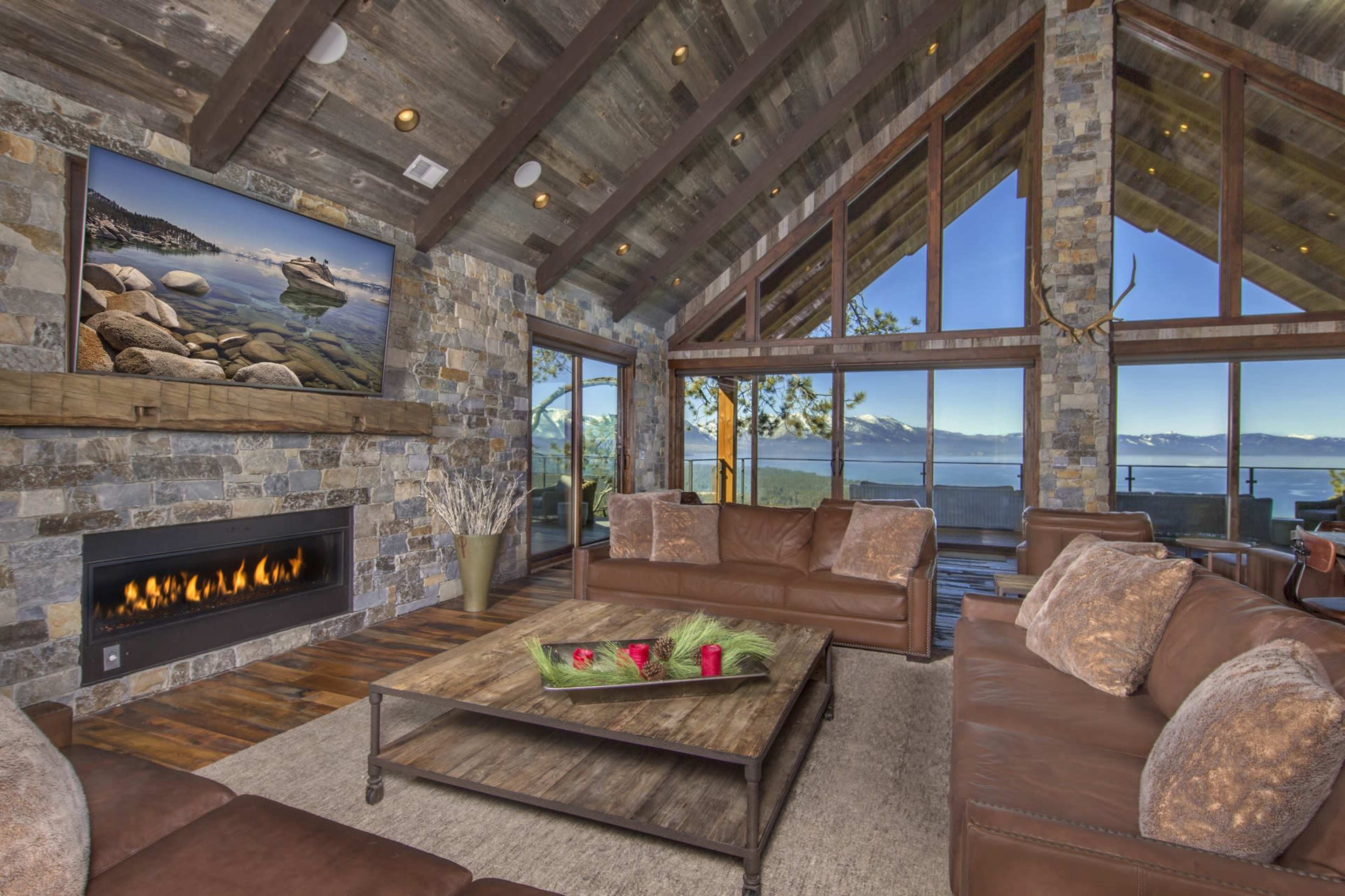The living room features a stone fireplace, large windows with a view of the mountains, and leather furniture arranged around a coffee table.