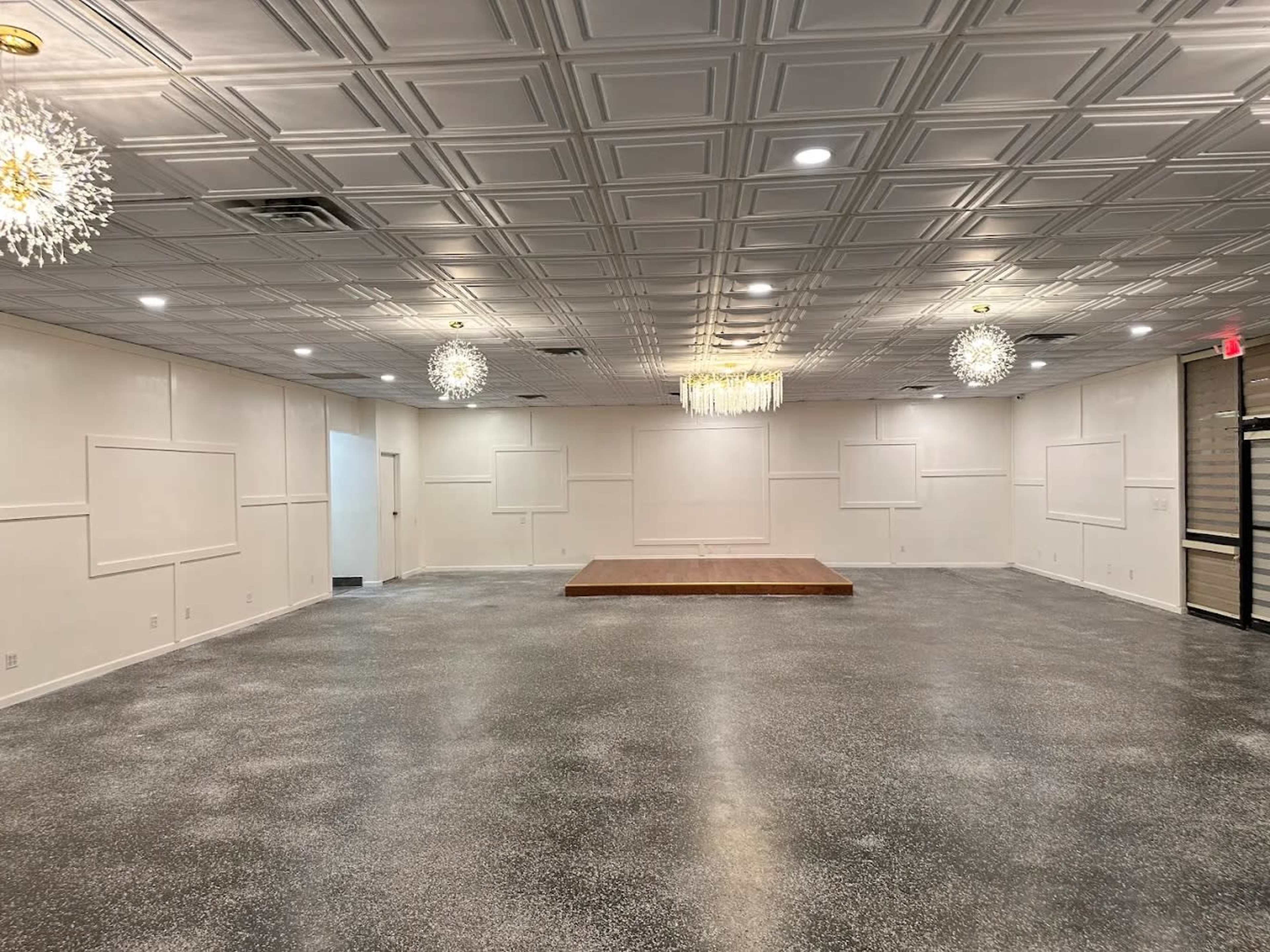 The image shows a spacious, empty room with a polished floor, high ceiling, and a small raised wooden stage at the center, illuminated by decorative light fixtures.