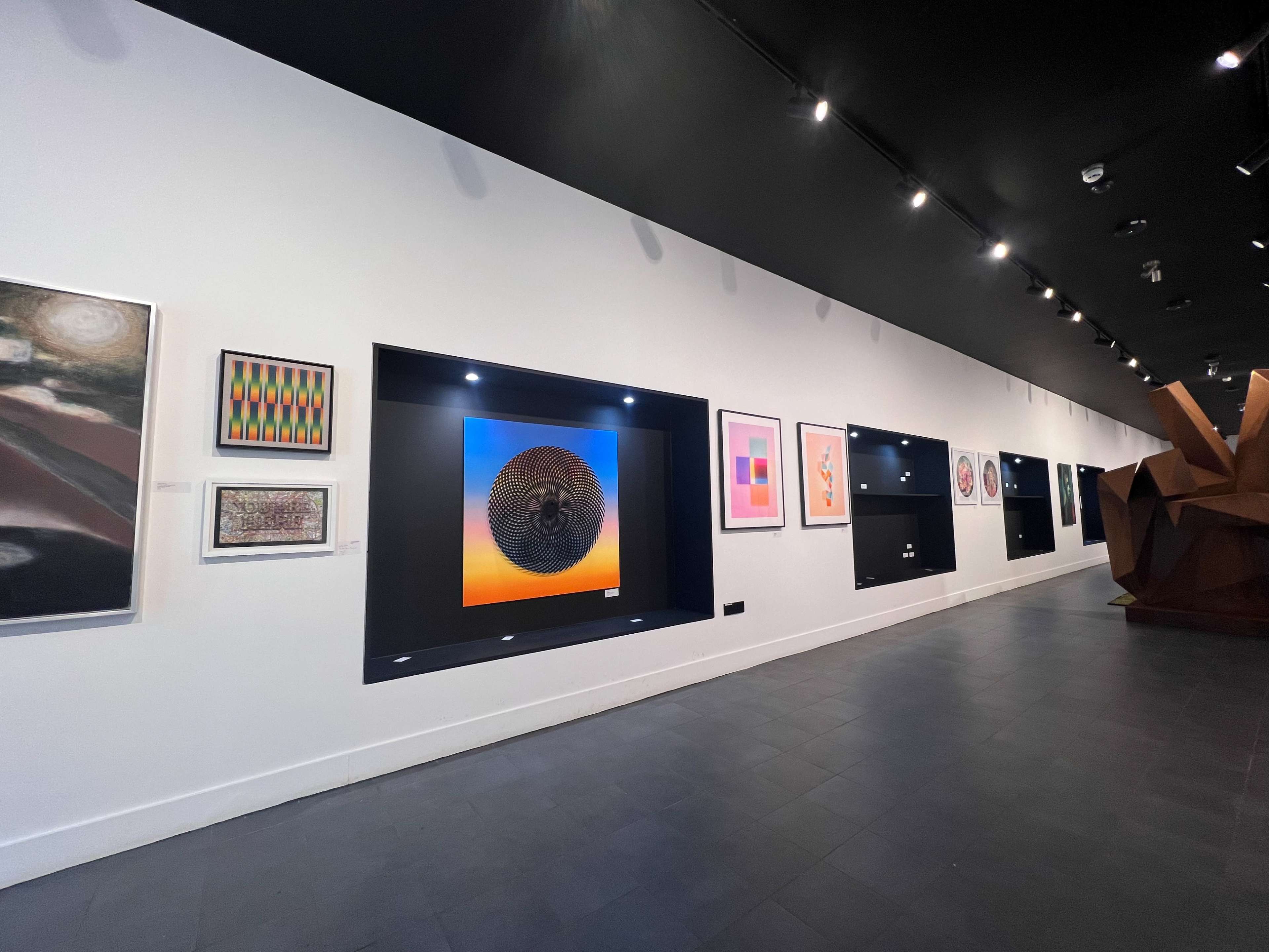 An interior of an art gallery with various framed artworks displayed on a white wall and a large sculptural piece on the right.