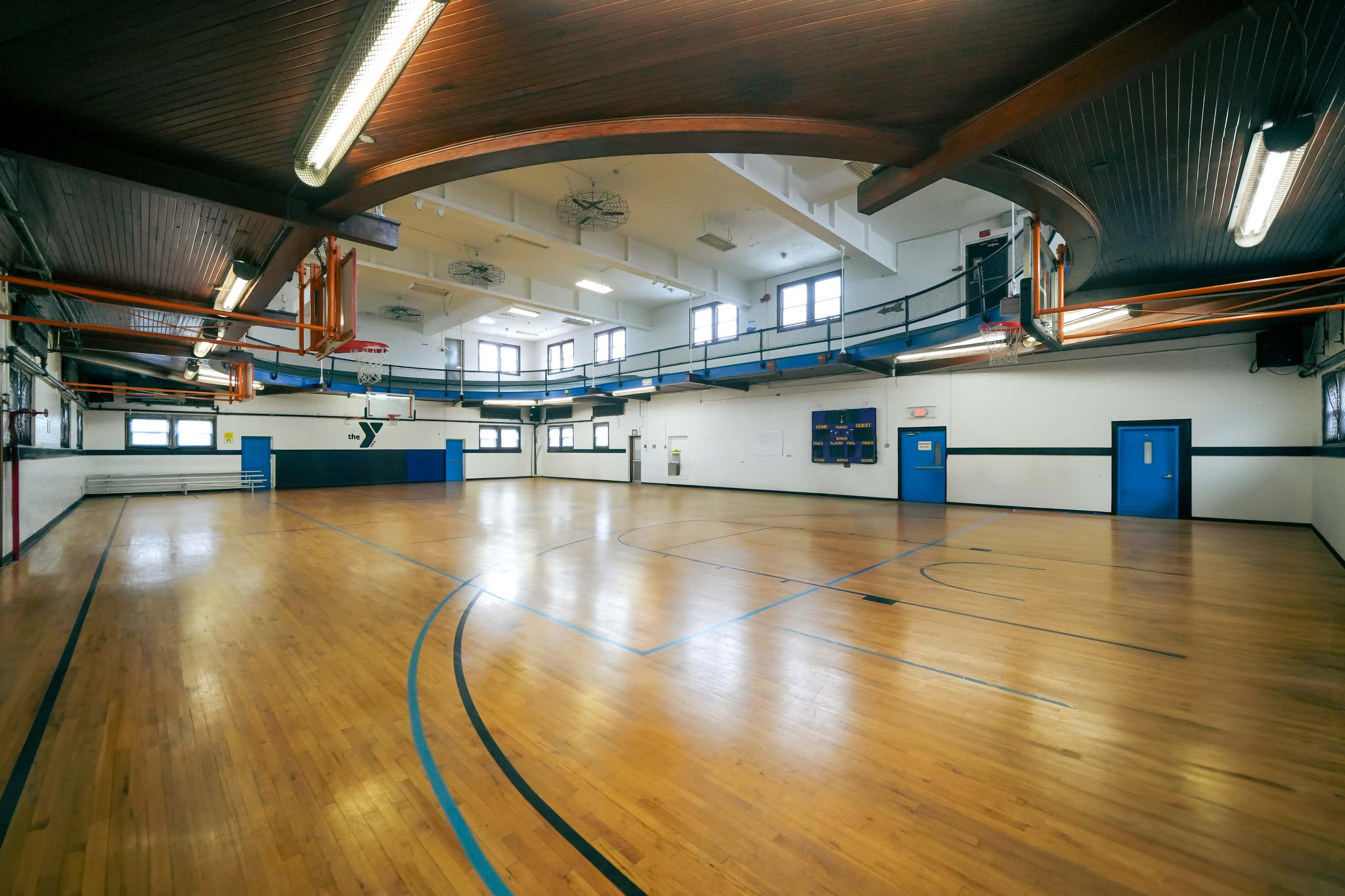 The image shows a spacious gymnasium with wooden floors, overhead basketball hoops, and blue and white walls.