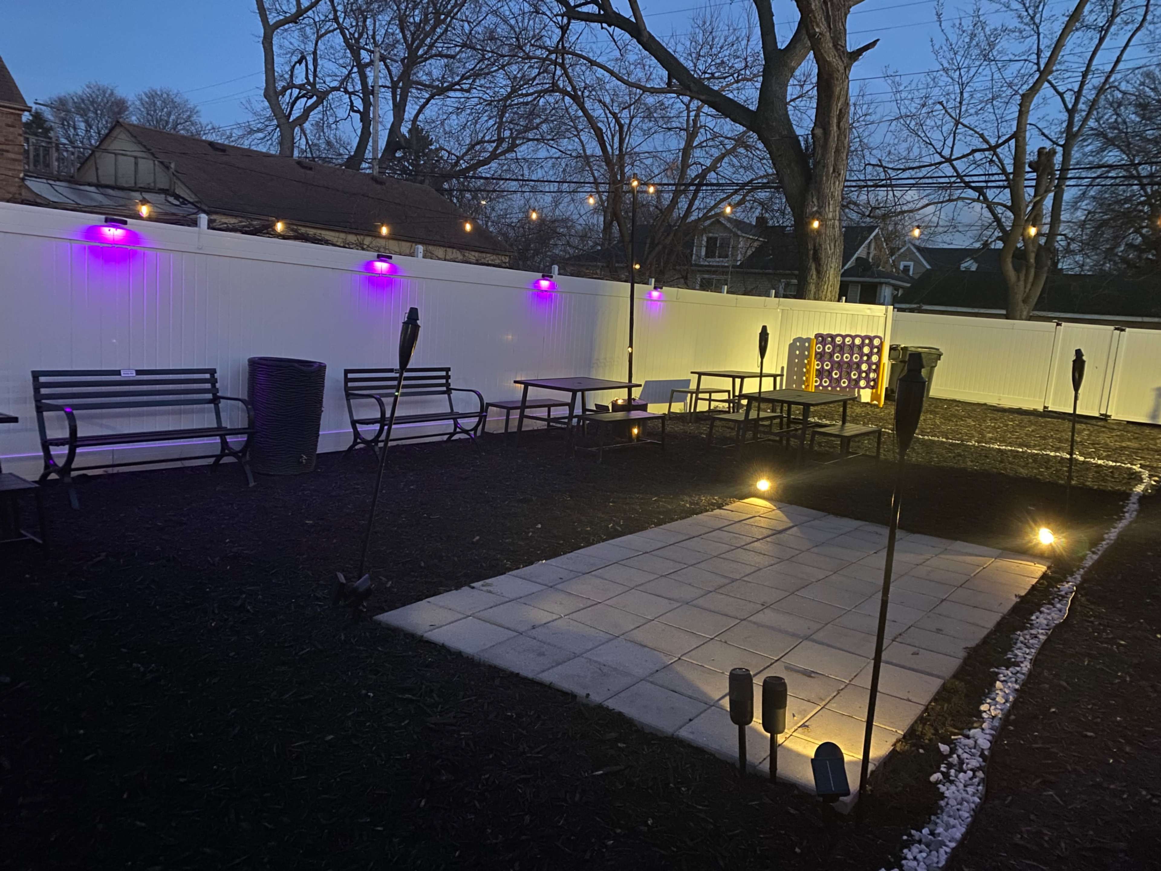 The image shows an outdoor patio area with purple lights, seating arrangements, and a paved square surrounded by tiki torches.