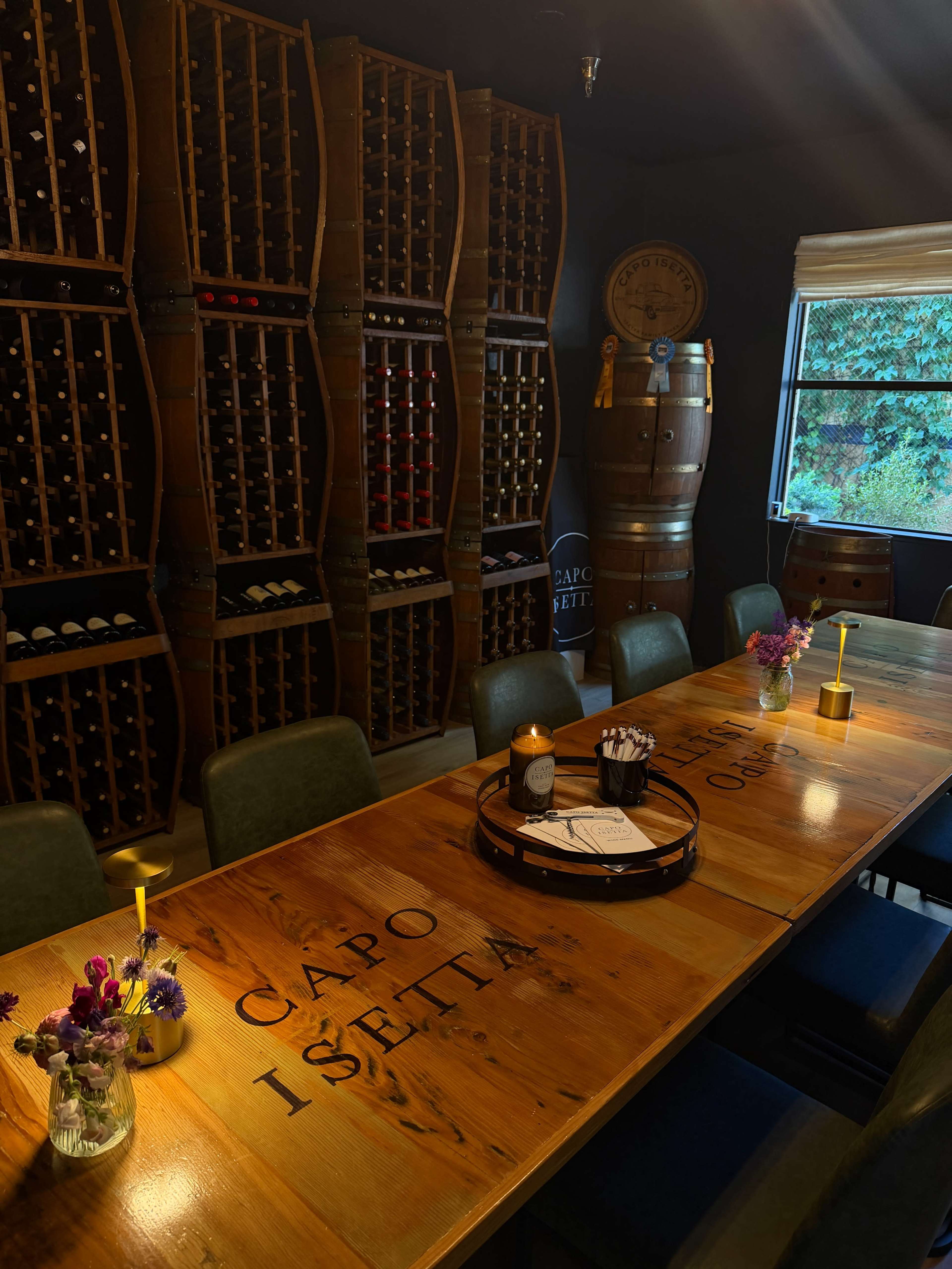 The image shows a dimly lit wine cellar with a wooden table, surrounded by chairs, and a wall of wine racks filled with bottles.
