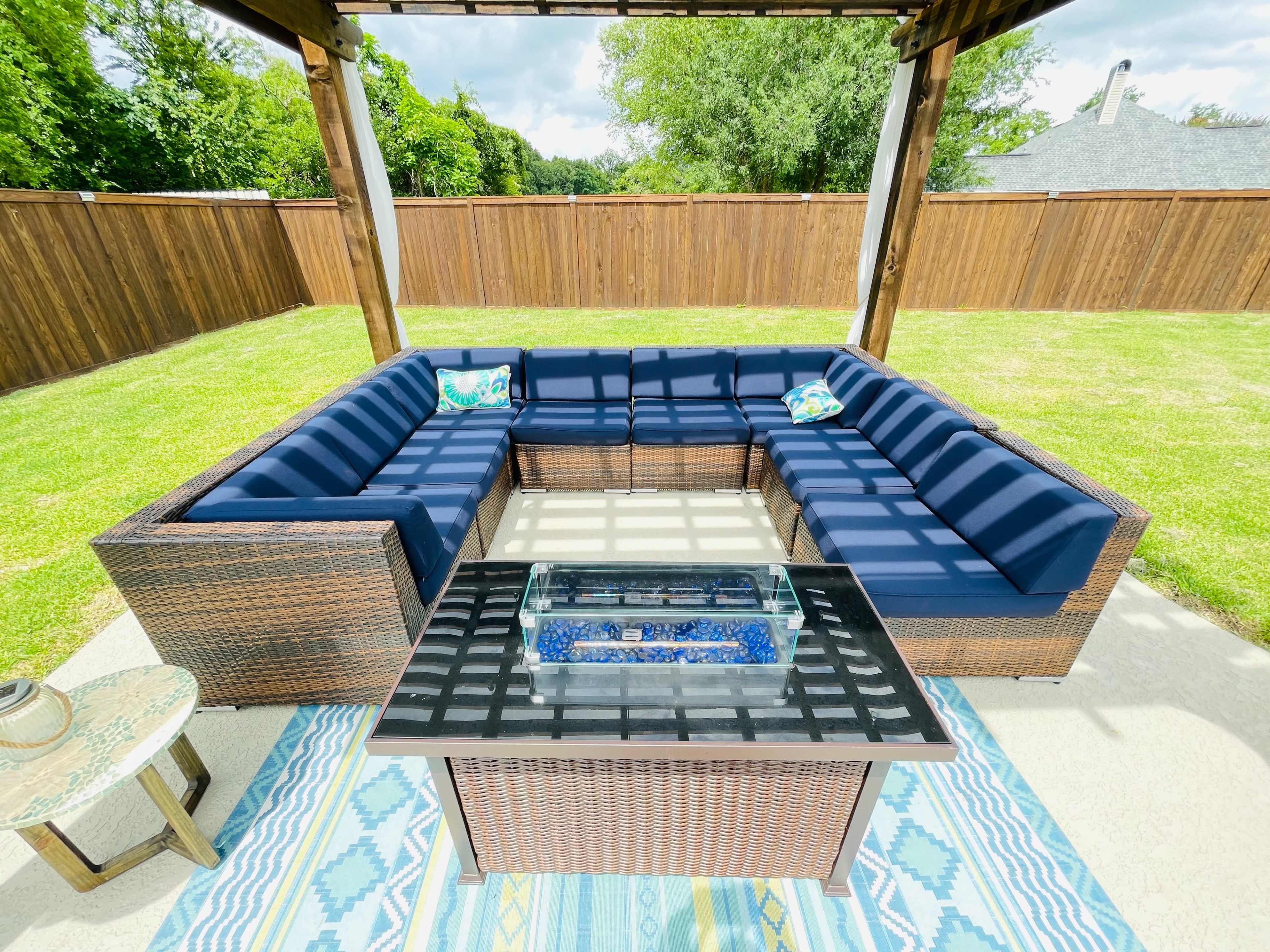 A spacious outdoor seating area features a large sectional couch with navy cushions and a glass-top fire pit table, all set on a patio surrounded by a grassy yard and wooden privacy fence.