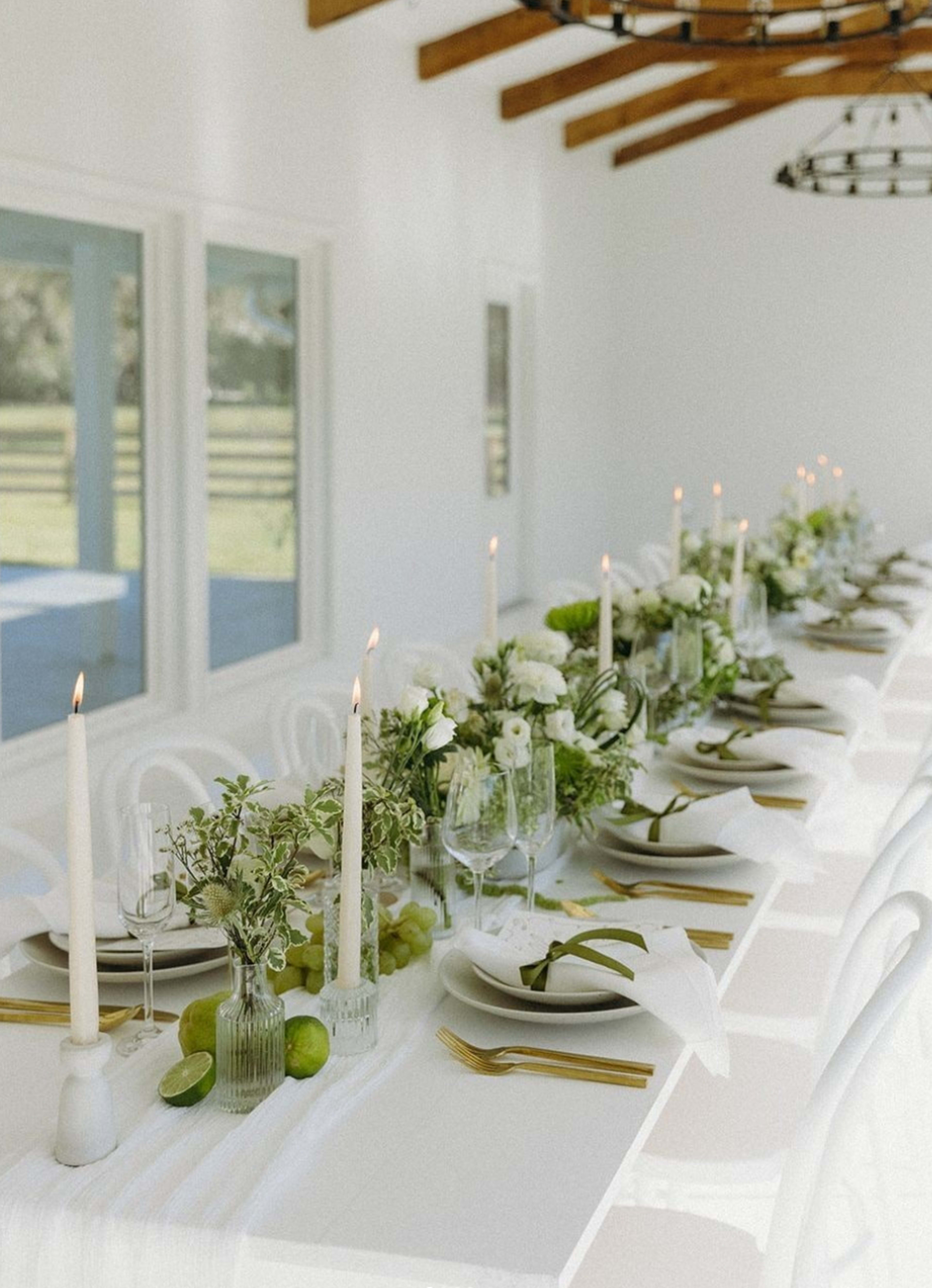 A long, white dining table is set with elegant tableware, flickering candles, and fresh floral arrangements alongside green limes.