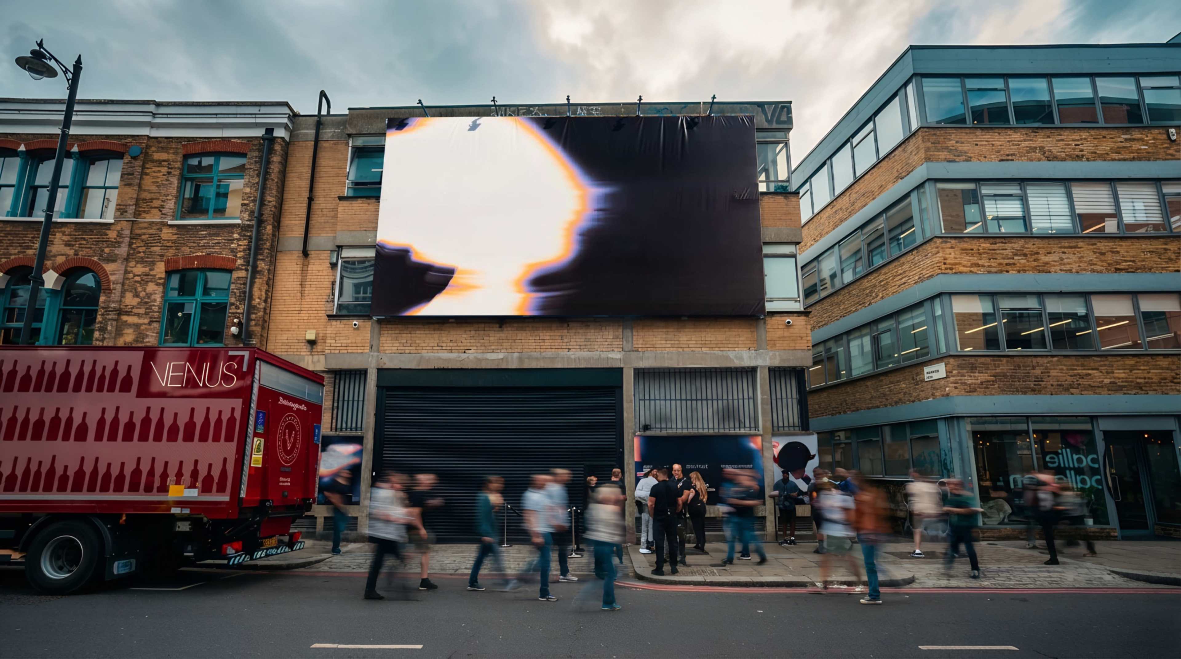 A large digital billboard displaying abstract imagery hangs on the side of a brick building as pedestrians walk by in a bustling urban area.