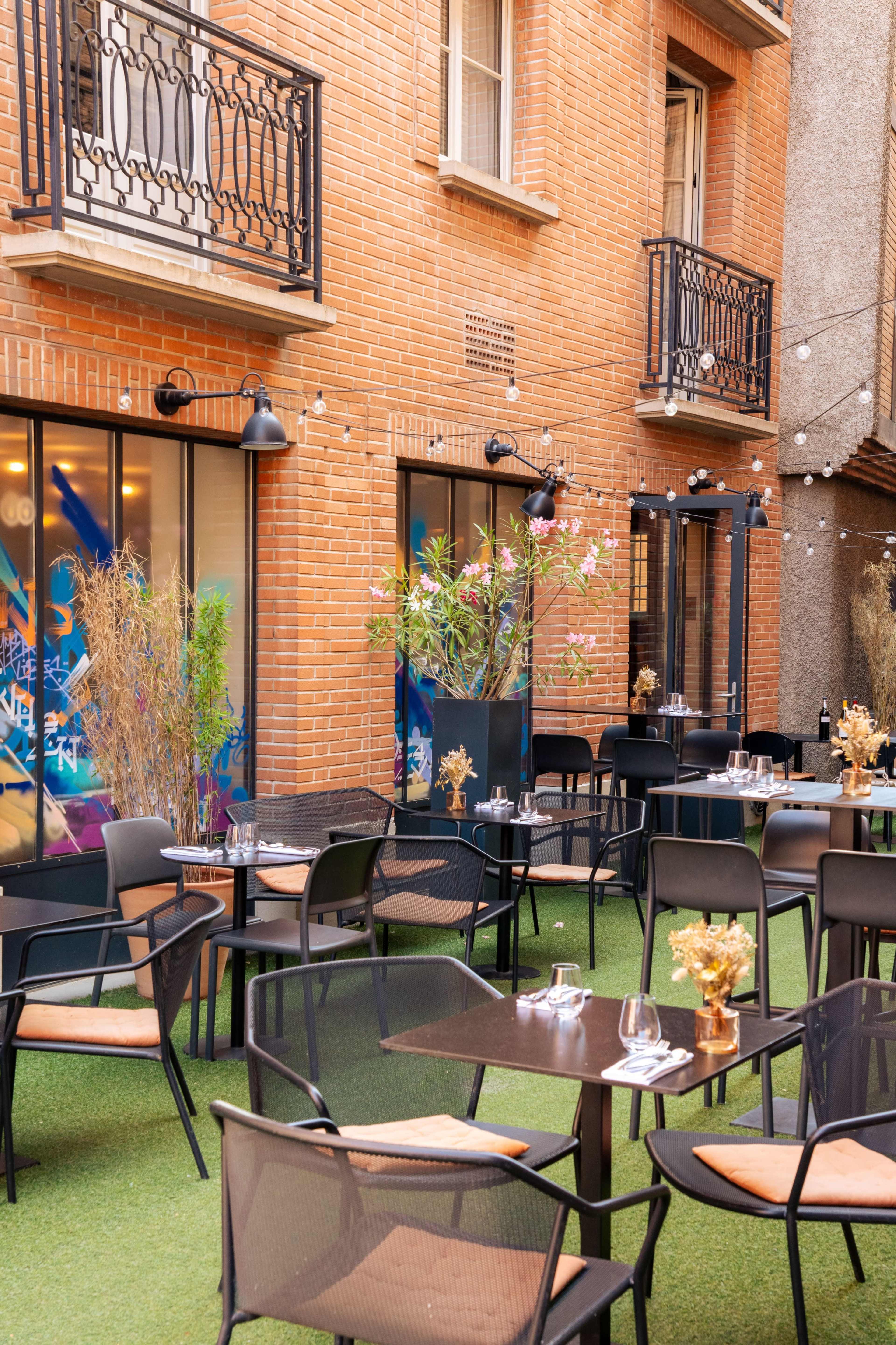 The image shows an outdoor dining area with black chairs and tables set up on green turf, surrounded by brick walls and decorative plants.