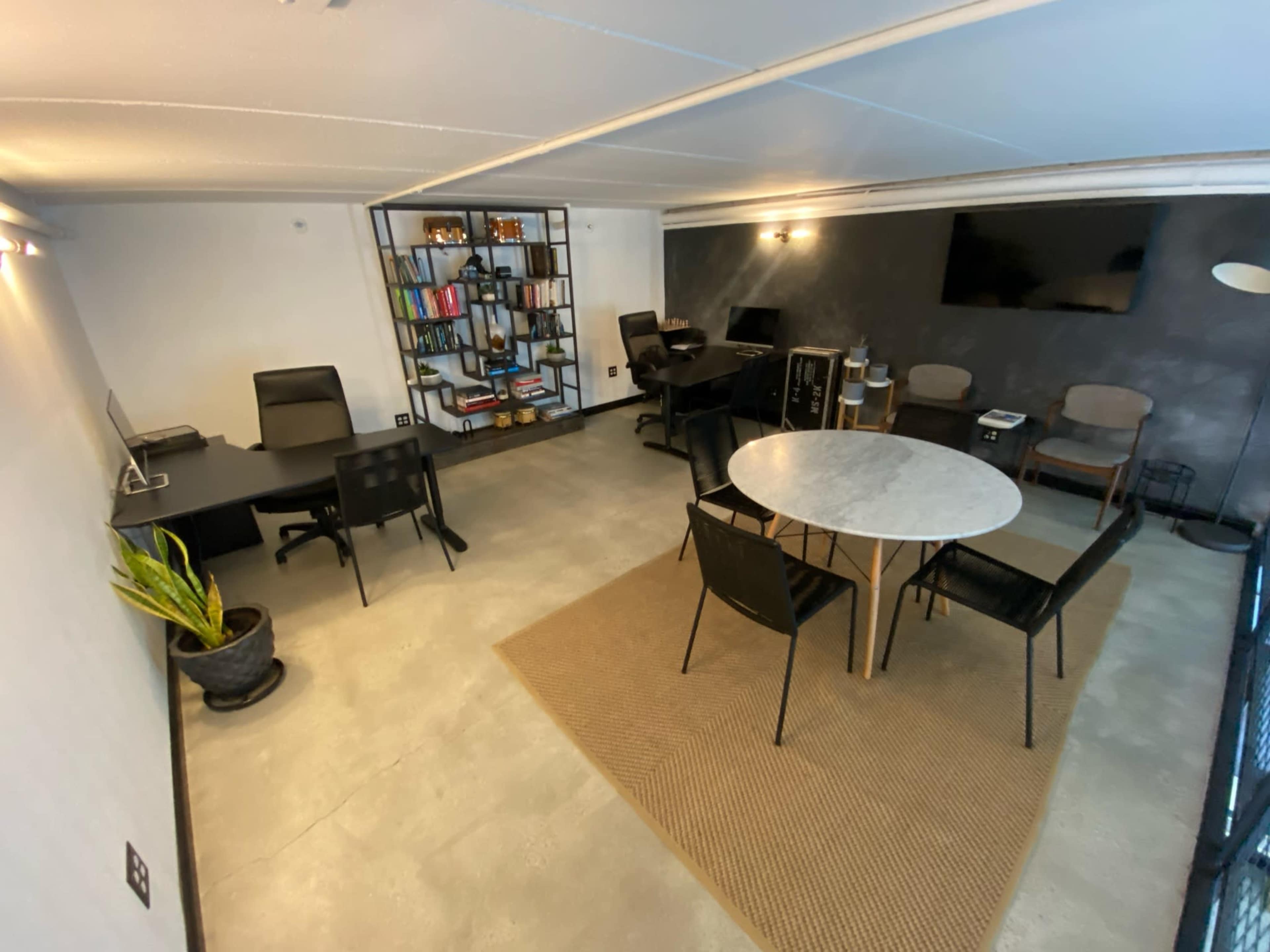 A modern office space featuring two desks with chairs, a round table with chairs, a bookshelf filled with books, and a wall-mounted television.