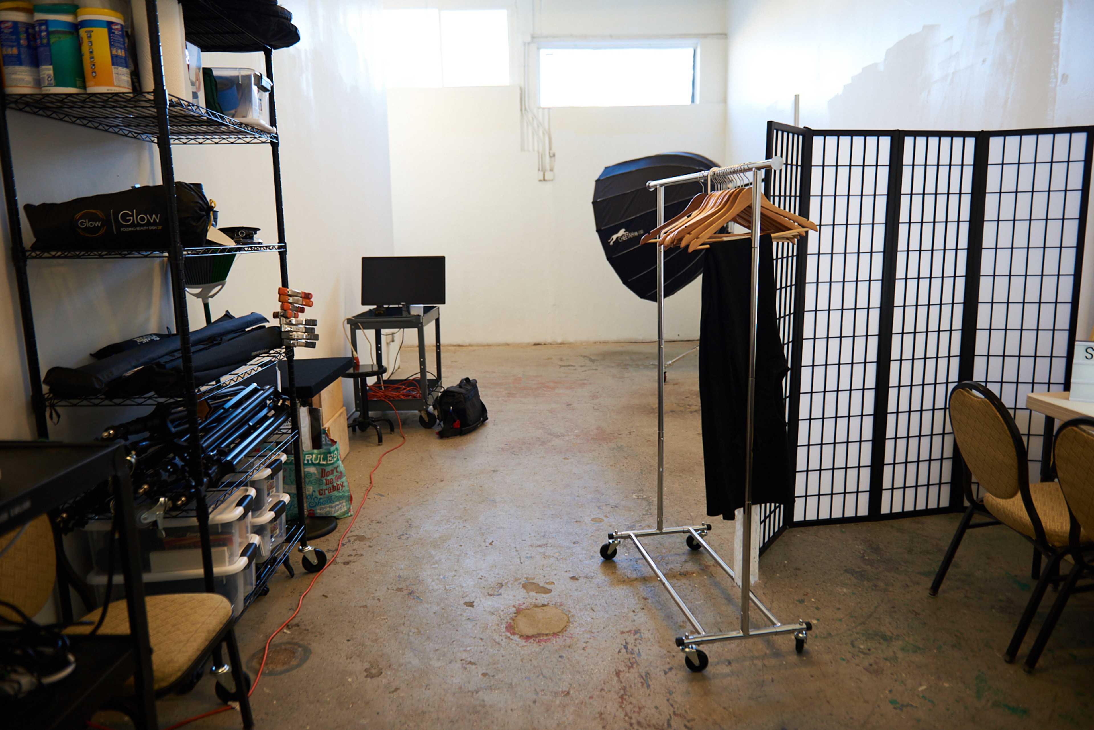 An empty studio space features a clothing rack, a monitor on a table, folding chairs, and a room divider.
