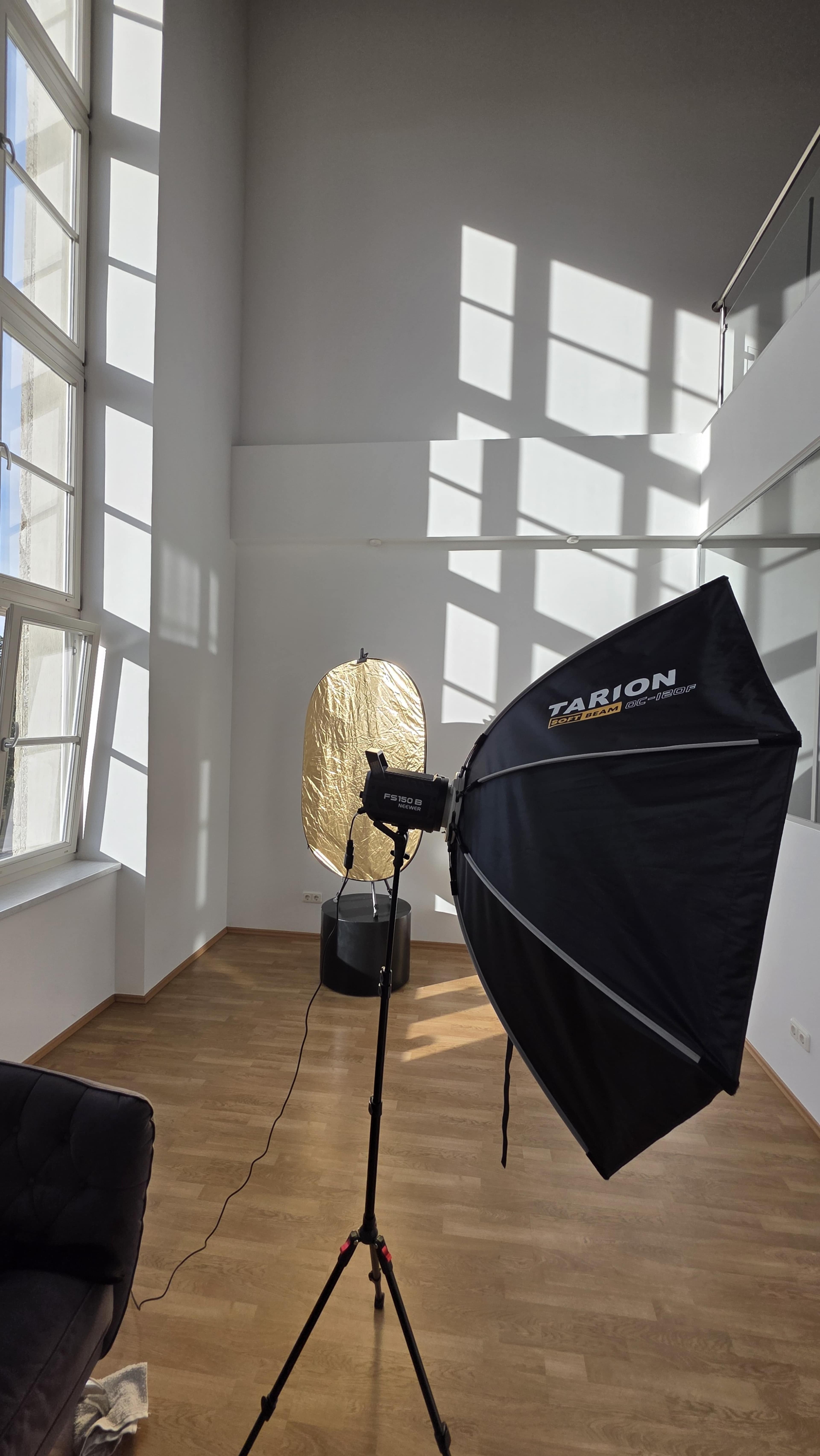 The image shows a bright room with large windows, featuring a photography setup that includes a softbox and a round reflector on a stand.