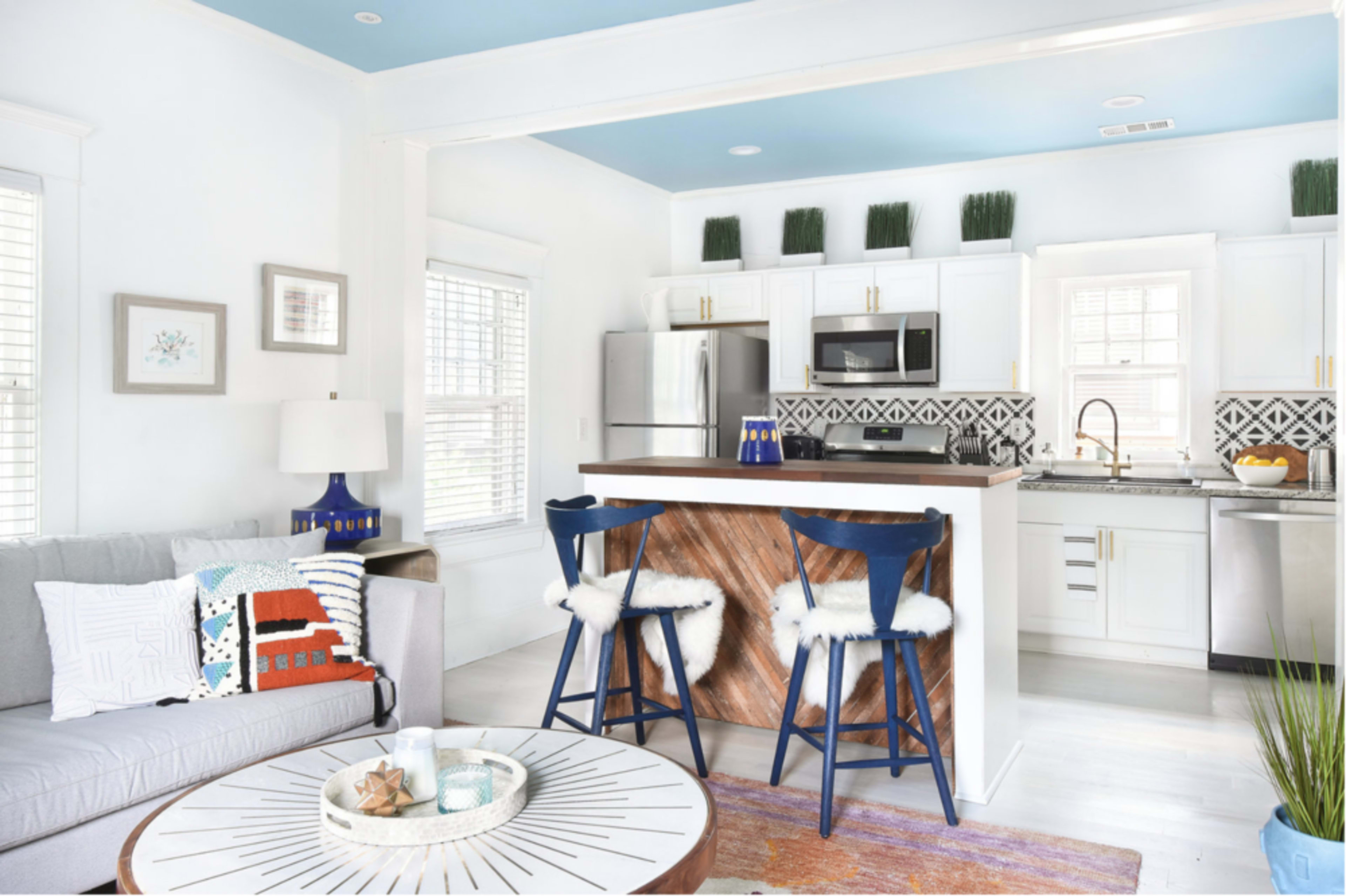 A modern kitchen and living area are connected, featuring a gray sofa, a round coffee table, and a kitchen island with blue stools.