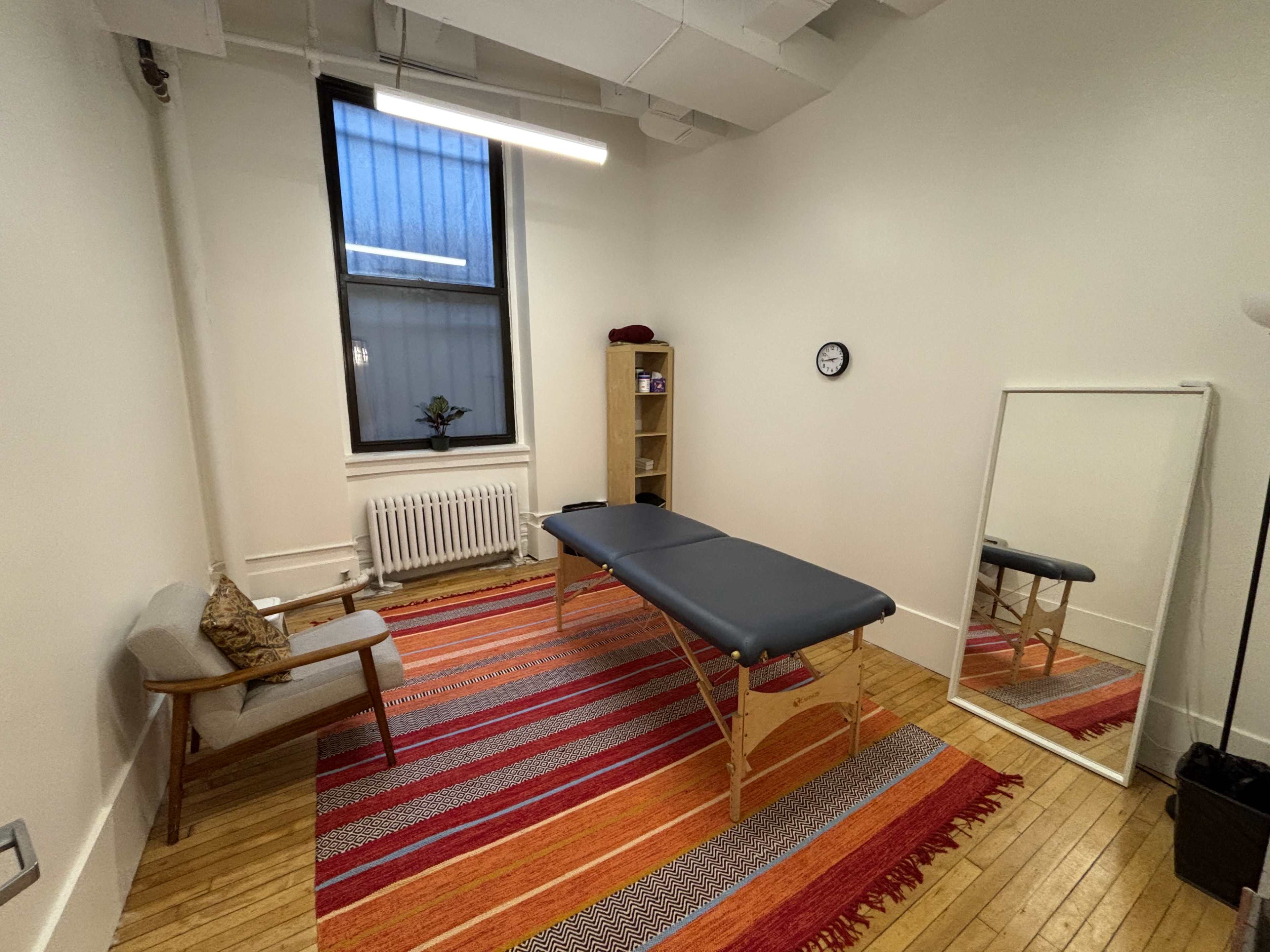 The image shows a well-lit therapy room featuring a treatment table, a chair, a mirror, and a shelf, all arranged around a colorful striped area rug.