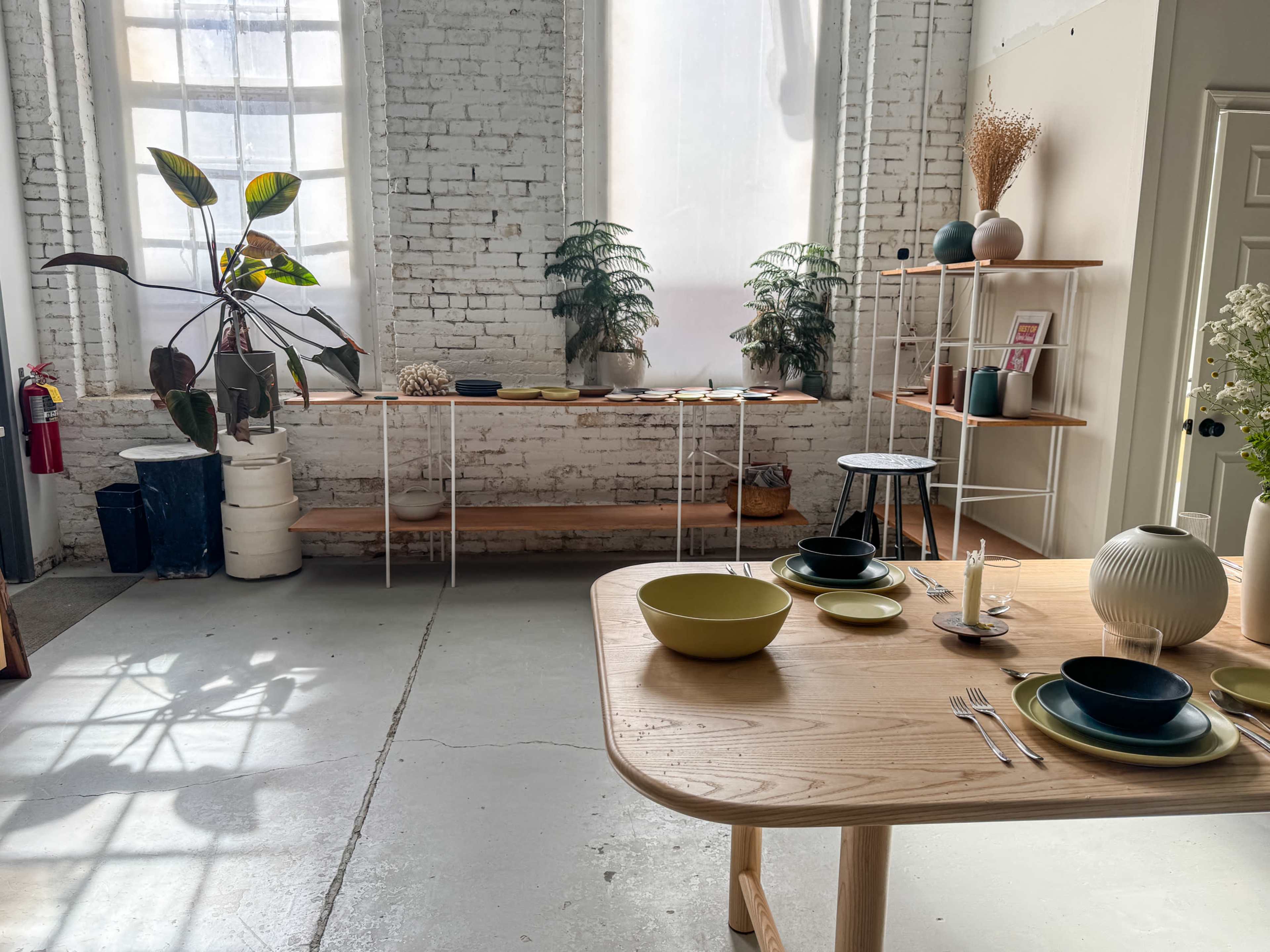 The image shows a bright interior space featuring a wooden table set with various dishes and utensils, surrounded by shelves displaying plants and decorative items against a backdrop of white brick walls and large windows.