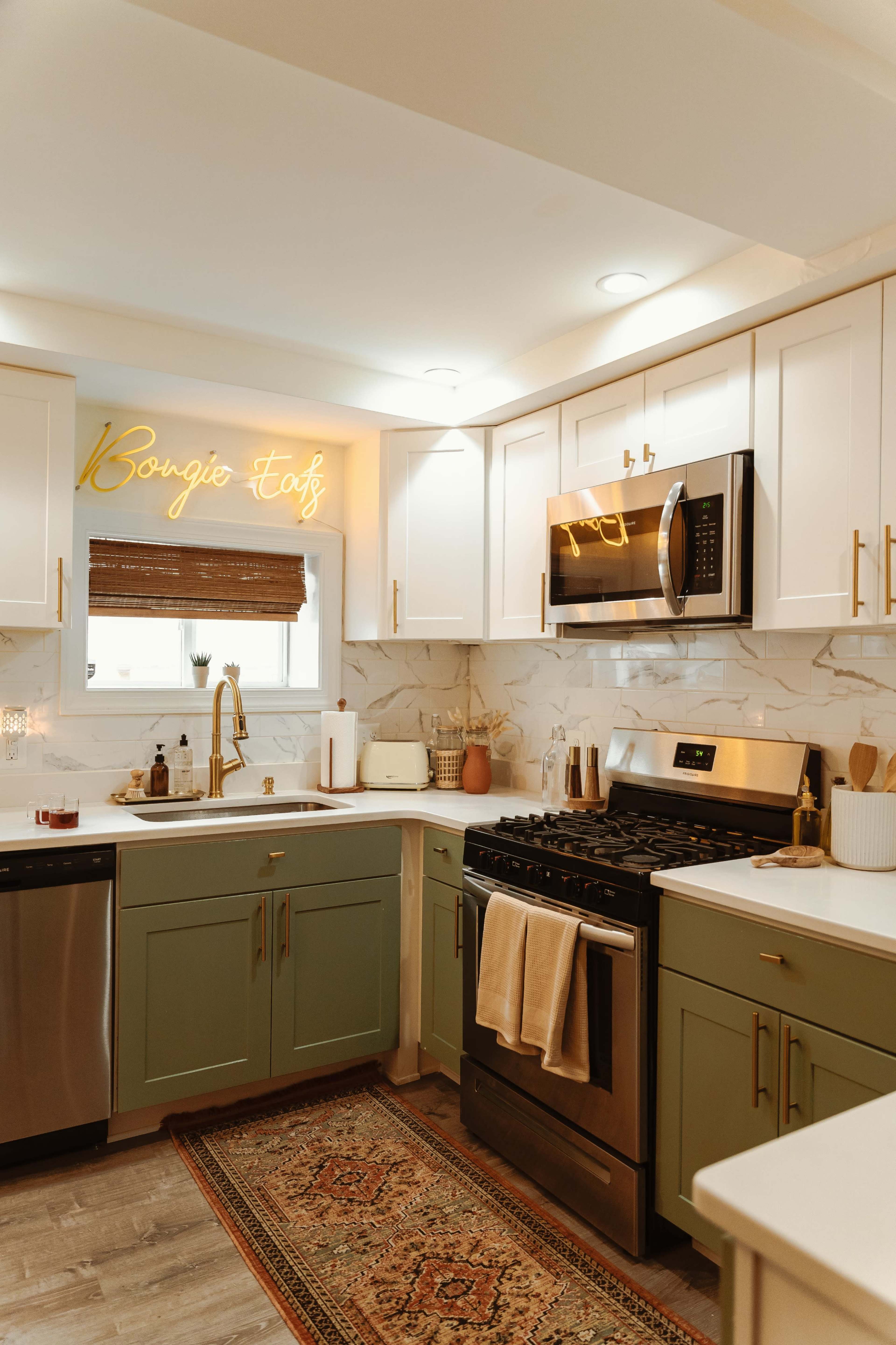 A modern kitchen features green and white cabinetry, stainless steel appliances, and a decorative rug, with warm lighting and a neon sign on the wall.