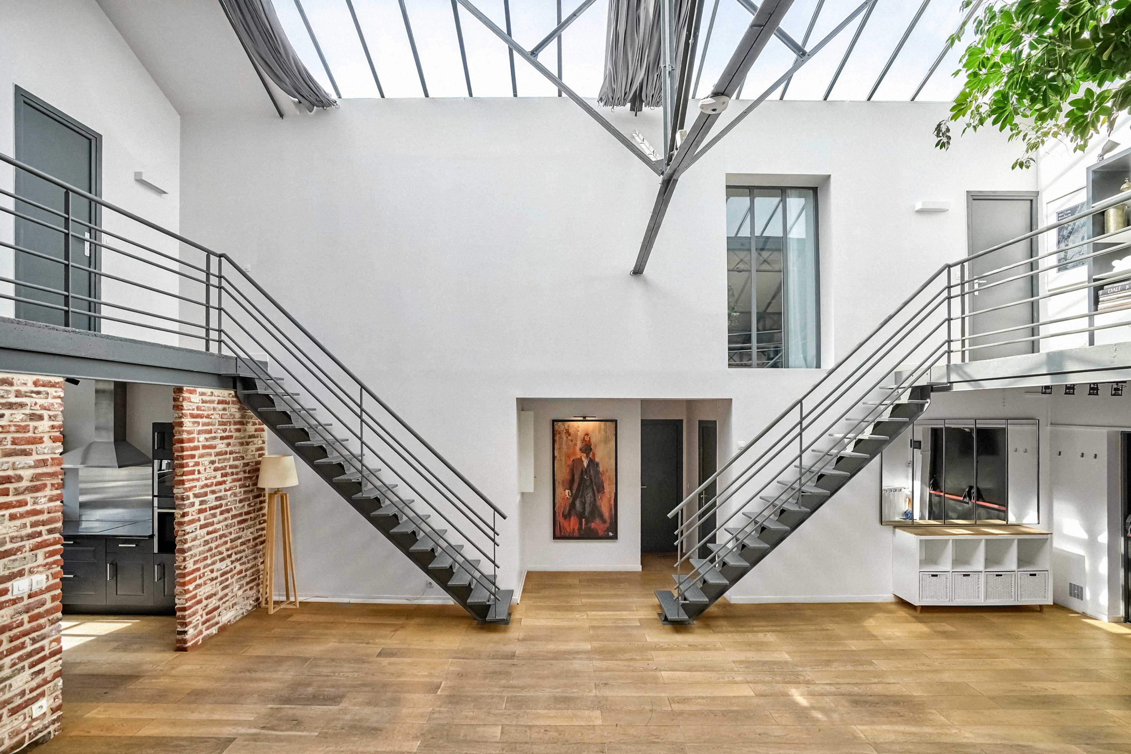 The image depicts a spacious interior with two metal staircases leading to an upper level, surrounded by white walls and exposed brick accents.