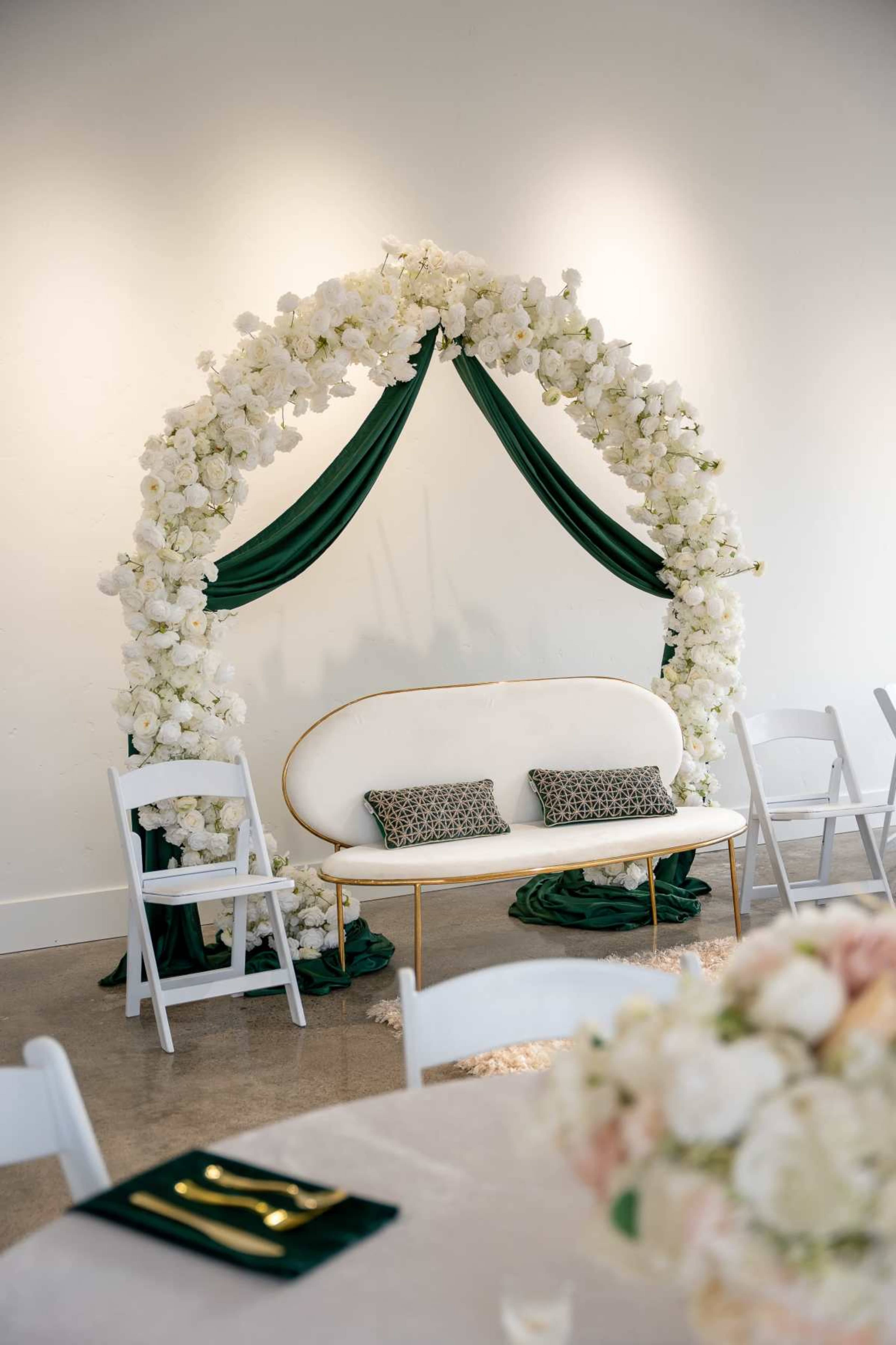 A floral arch with white flowers and green drapery surrounds a white sofa with two patterned cushions, set against a plain wall in a decorated space.