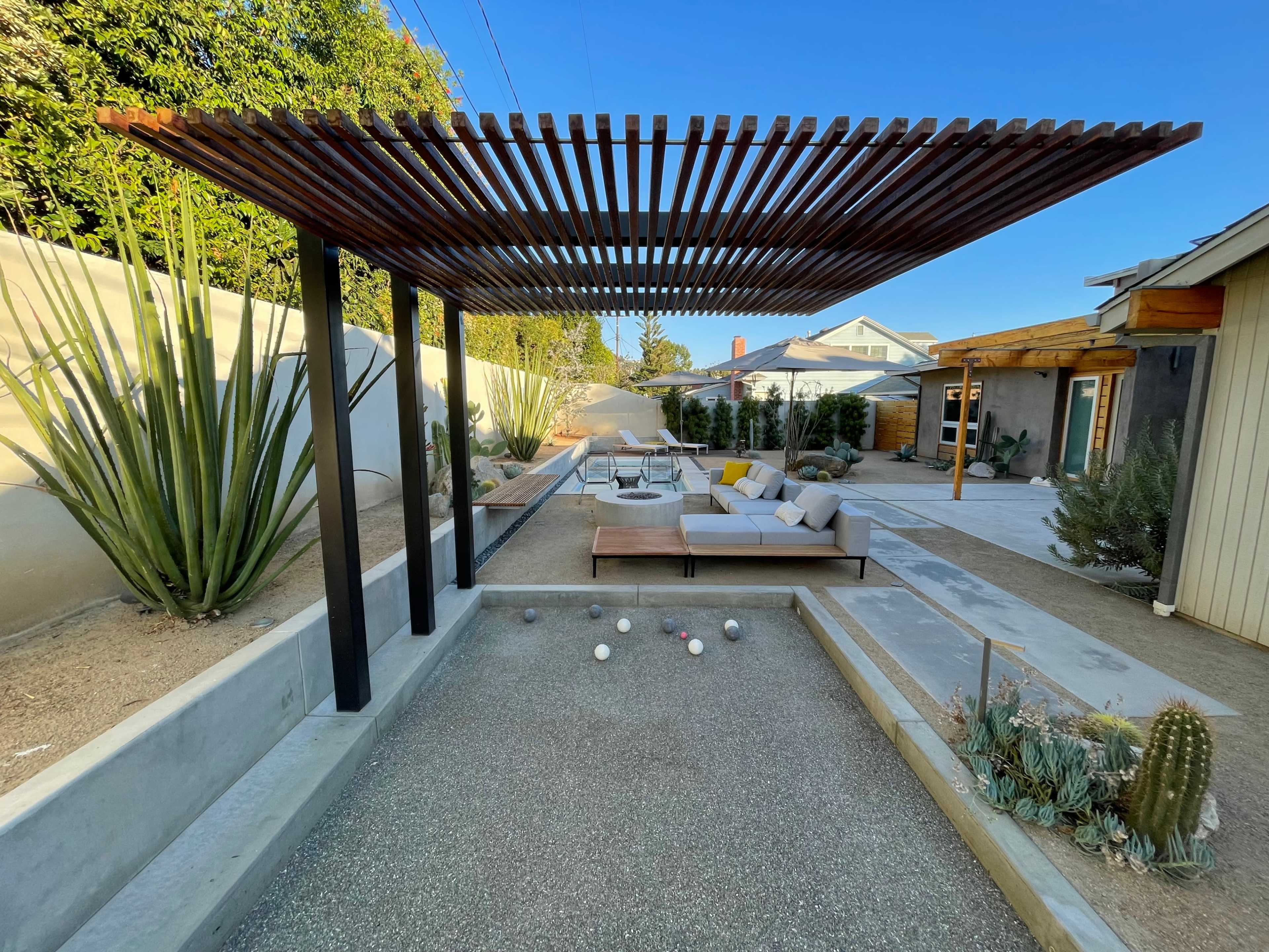 A modern outdoor space featuring a shaded seating area under a wooden trellis, with a gravel bocce ball court in the foreground and various desert plants surrounding the scene.