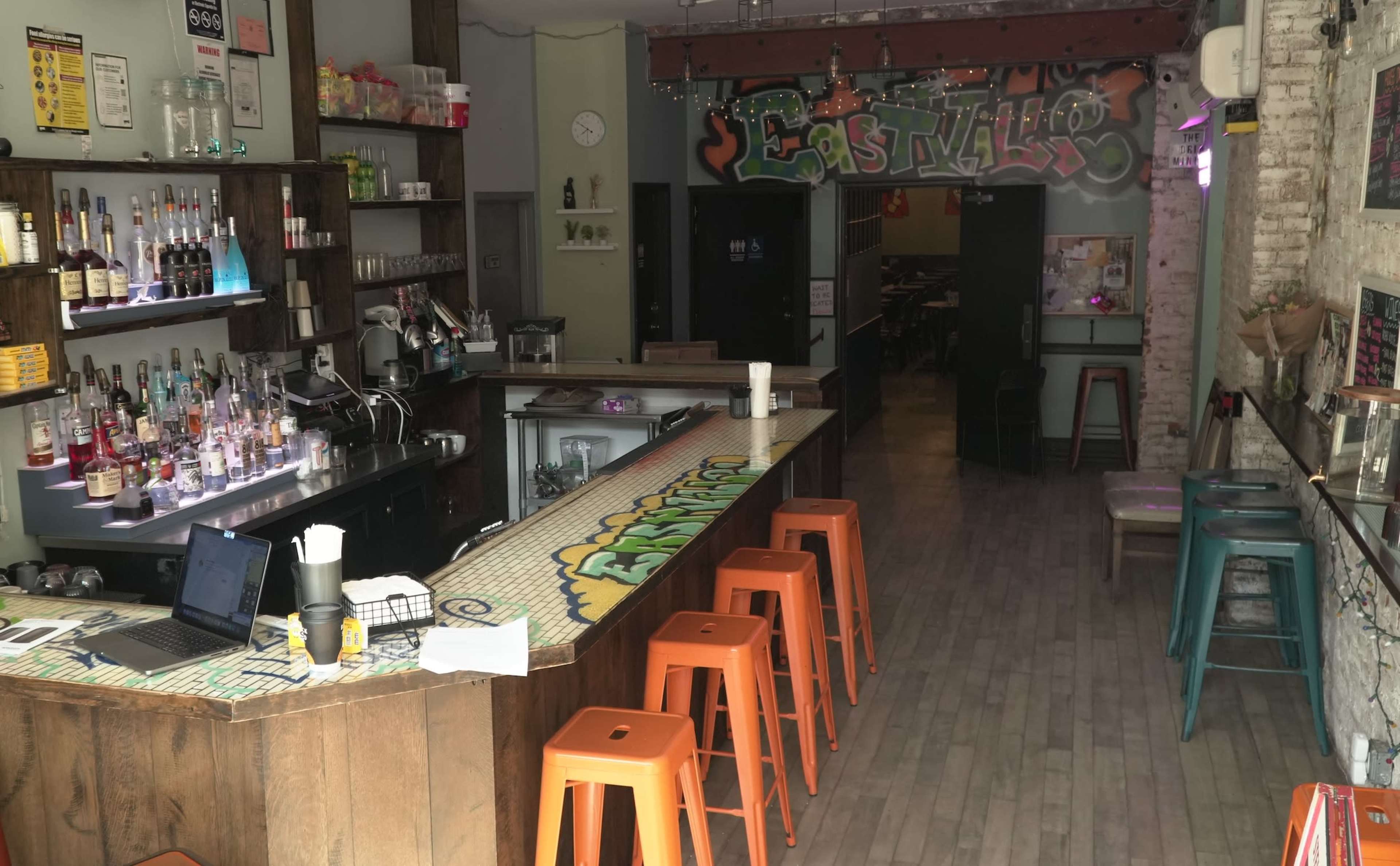 The image shows a bar area with colorful stools, a variety of liquor bottles on shelves, and a glimpse of a dining space in the background with graffiti art on the wall.