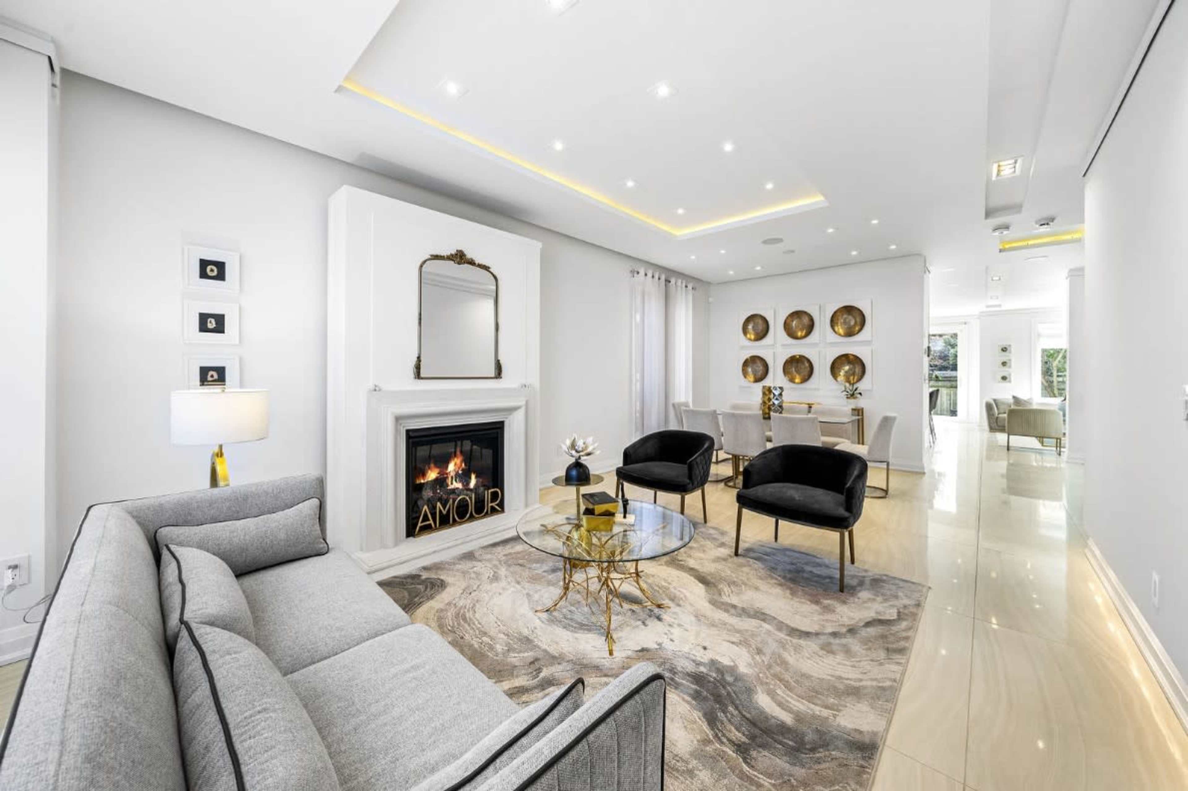 The image shows a modern living room with a gray sofa, two black chairs, a circular coffee table, and a fireplace framed by a mirror, accented by decorative wall art and natural light.