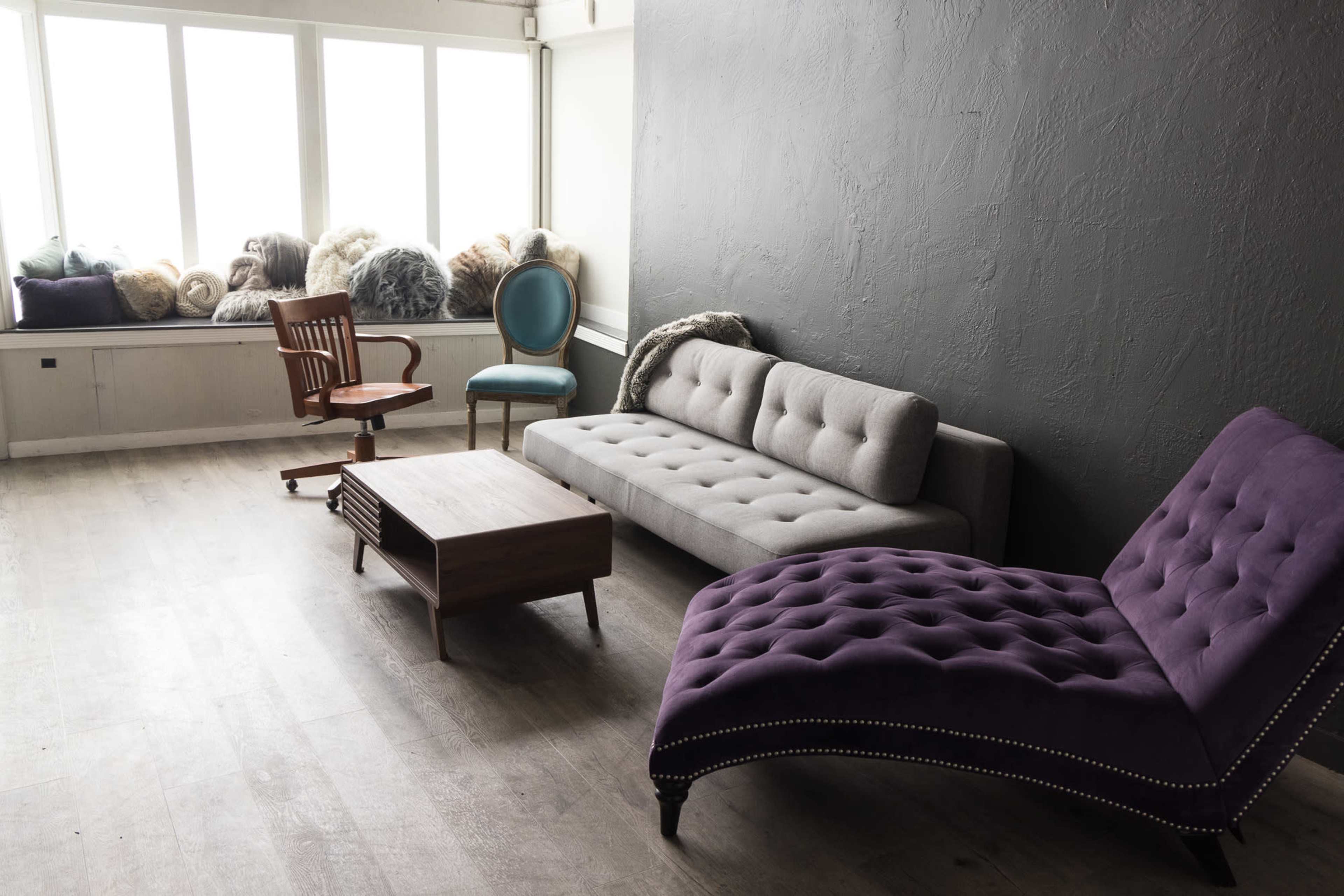 The image shows a minimalist room featuring a gray wall, a gray sofa, a purple chaise lounge, a wooden coffee table, and a wooden chair, with cushions placed on windowsills.