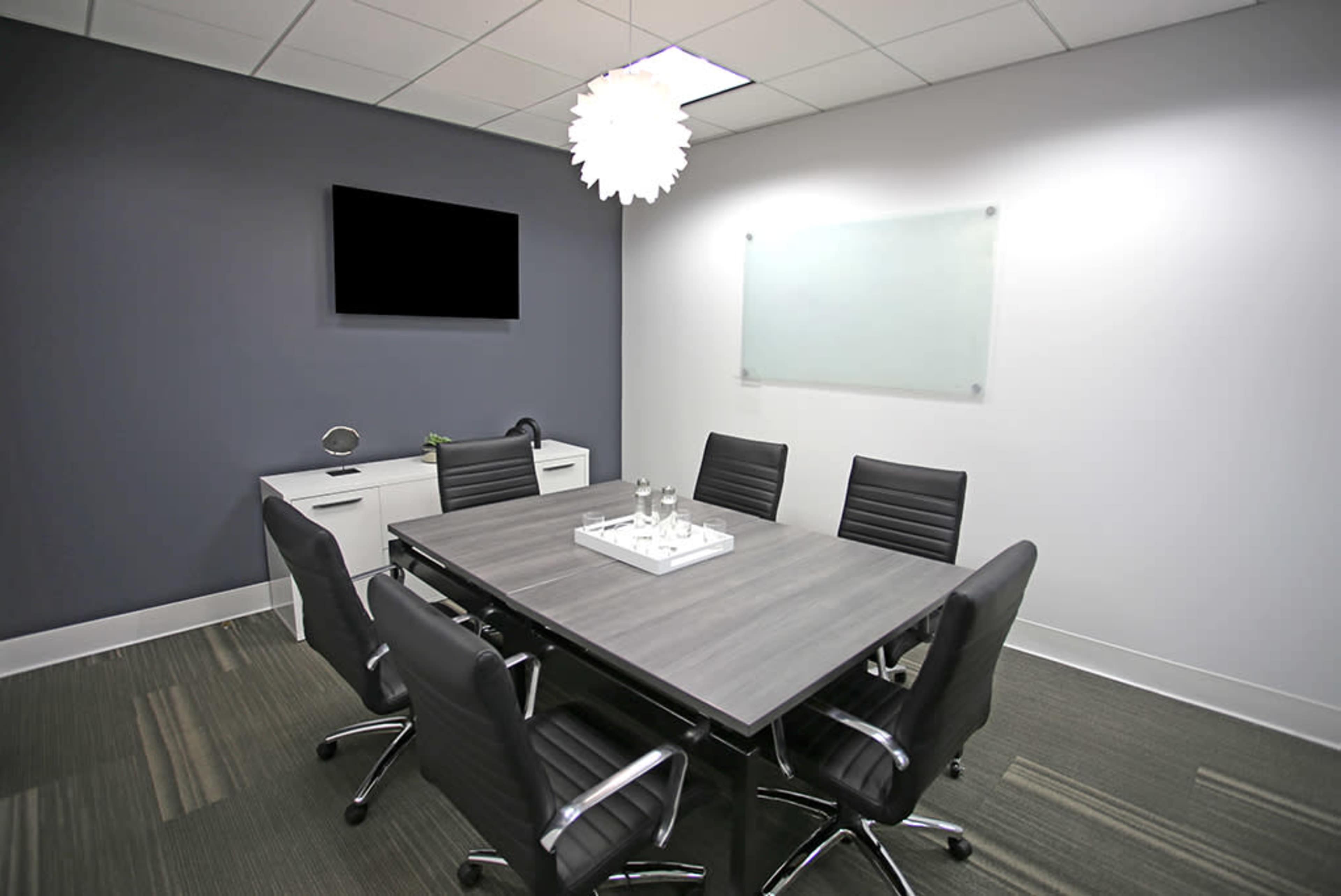A modern conference room features a rectangular table surrounded by black rolling chairs, a wall-mounted TV, and a whiteboard.