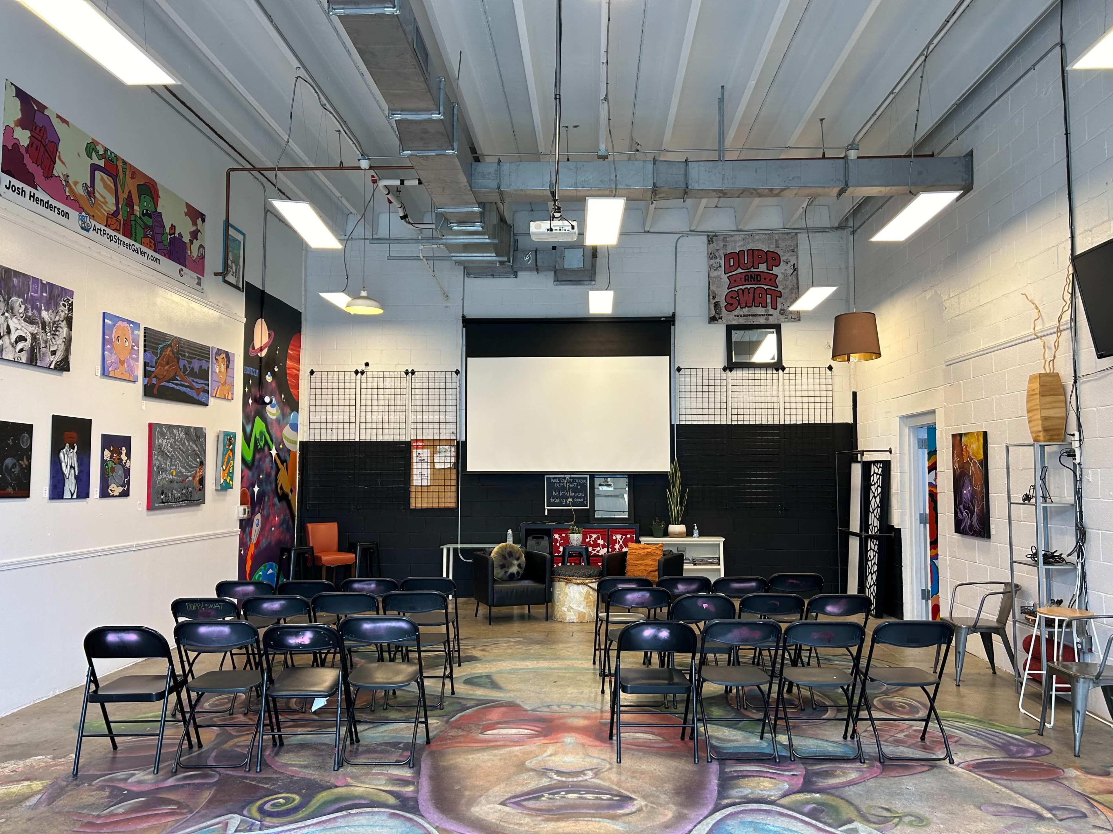 The scene shows a spacious room set up for a presentation, featuring several rows of chairs facing a large screen, with colorful artwork on the walls and decorative items around the space.