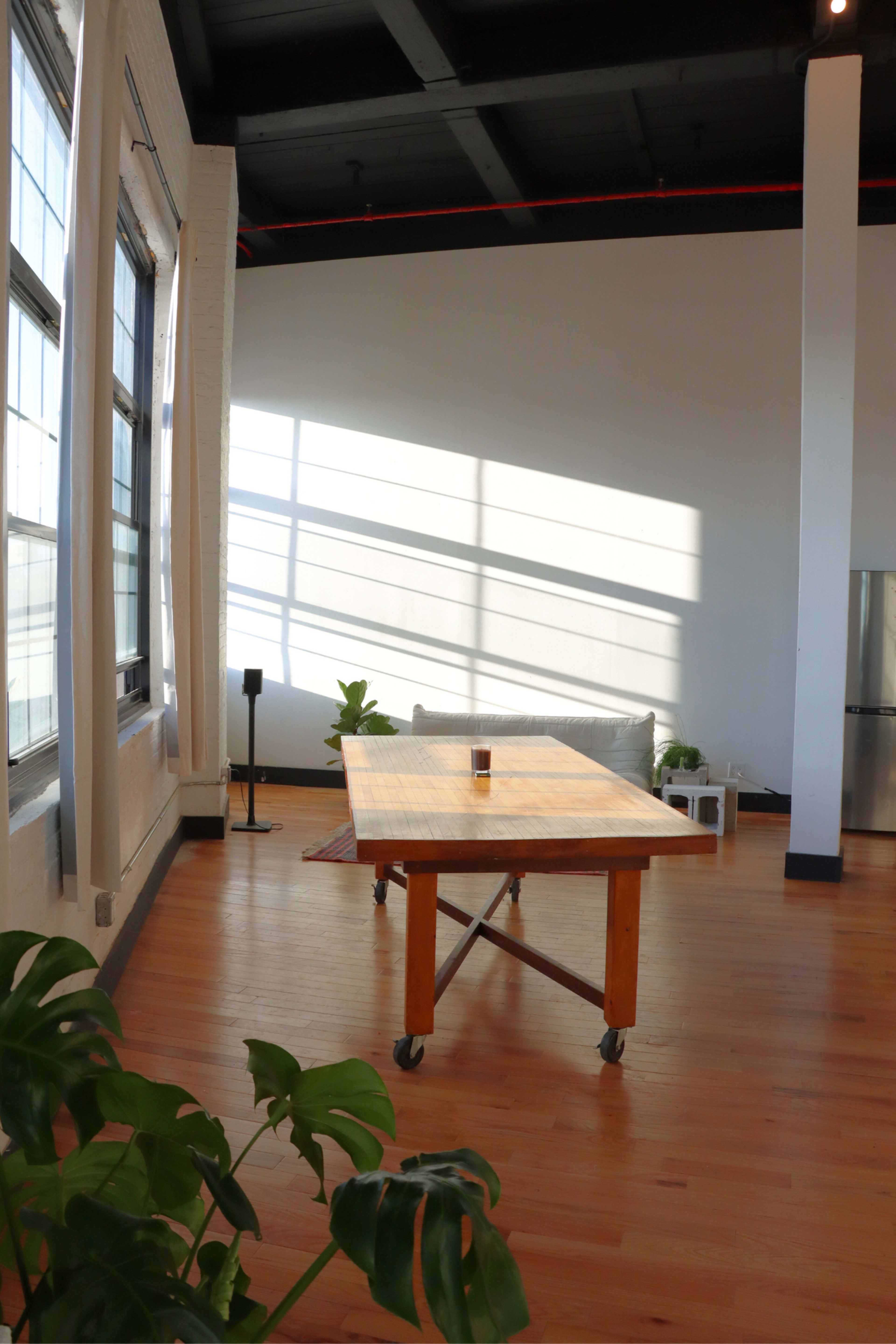 The image shows a spacious room with a wooden table on wheels, large windows casting shadows on the floor, and a few green plants.