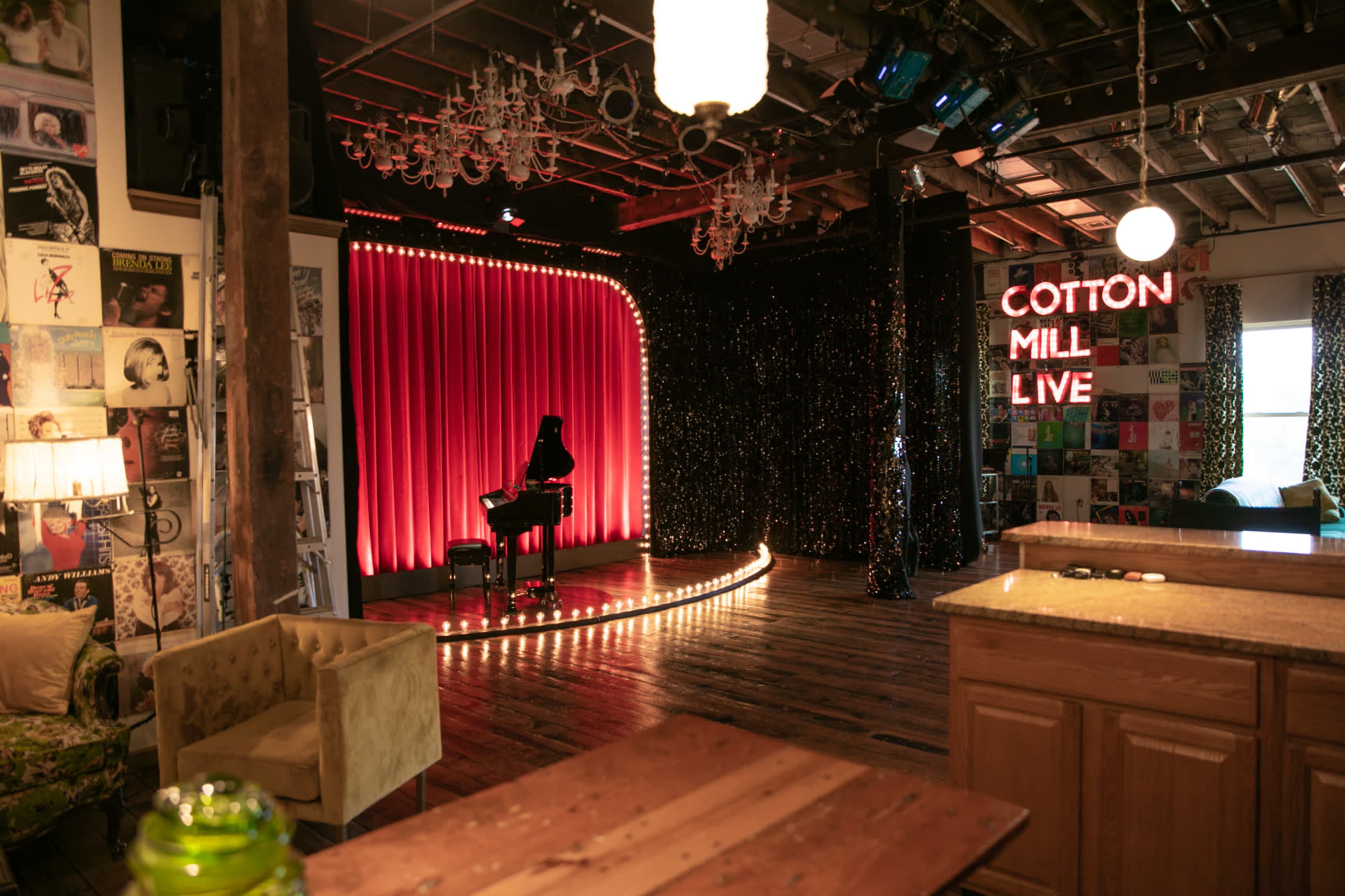 A performance stage at Cotton Mill Live, featuring a red curtain, a piano, and illuminated signage, with a cozy seating area visible in the foreground.