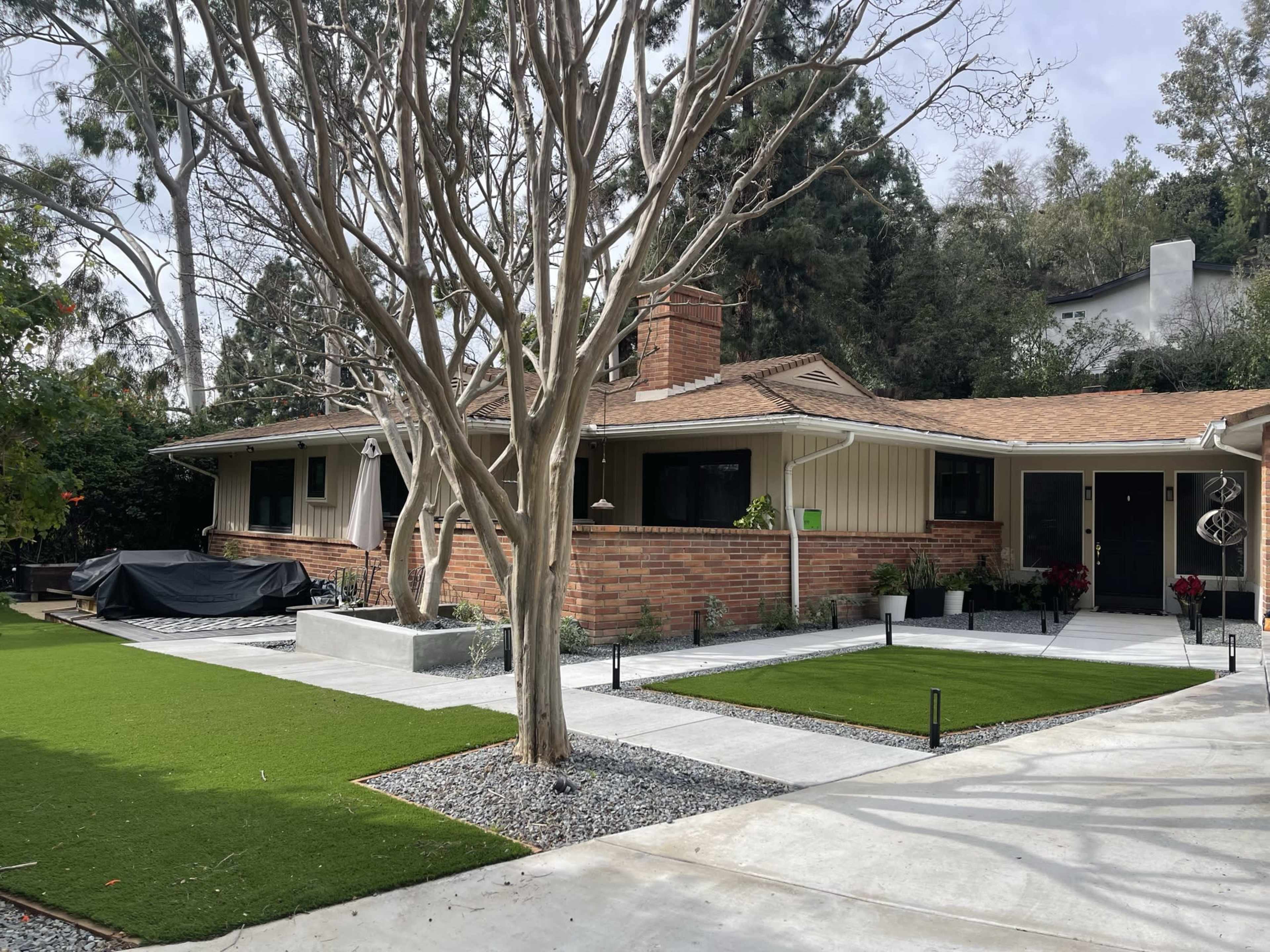 A single-story brick house with a landscaped front yard featuring artificial grass, gravel pathways, and trees.