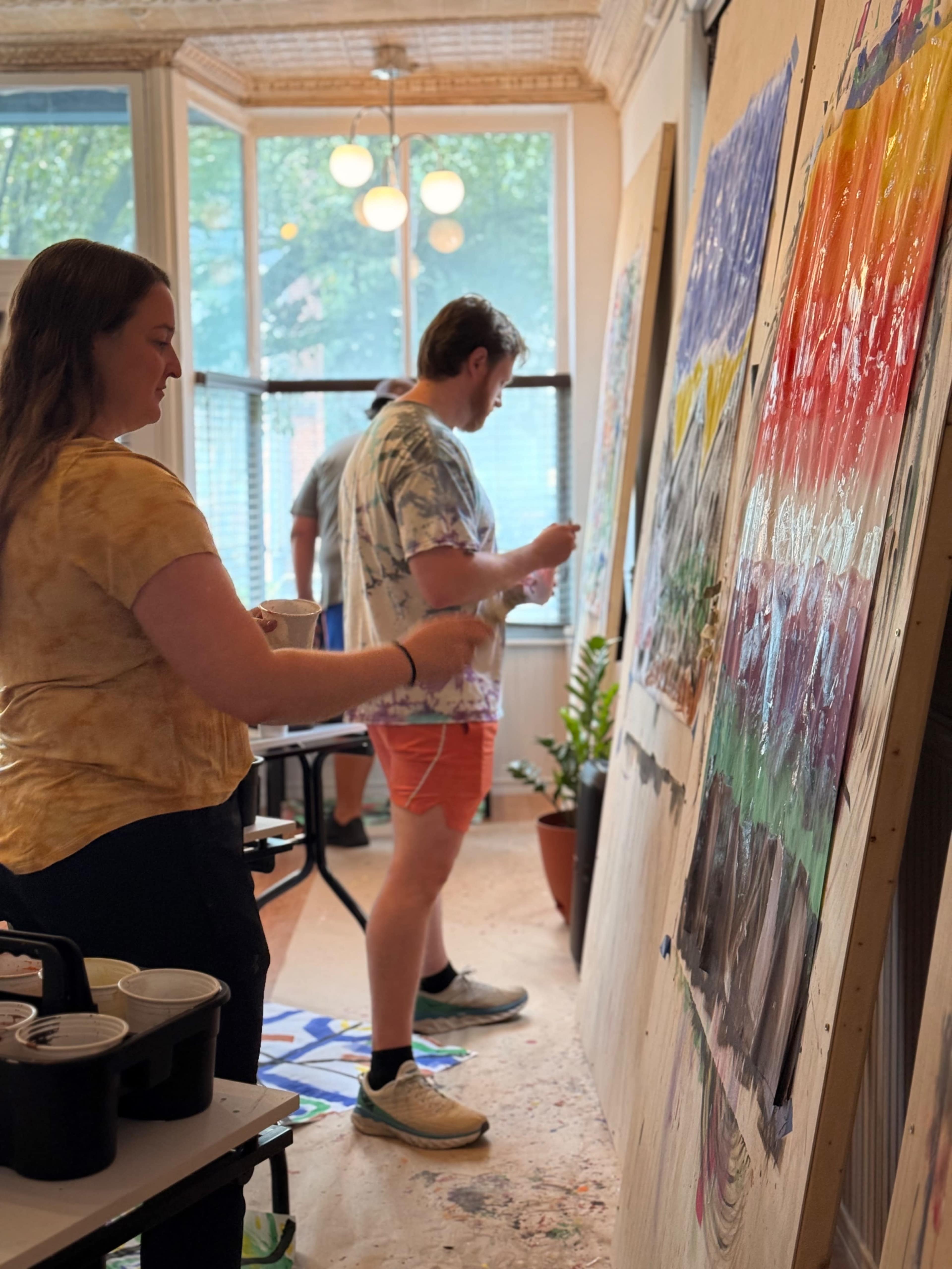 Two individuals are engaged in painting on a large wooden board in a brightly lit room filled with plants and natural light.