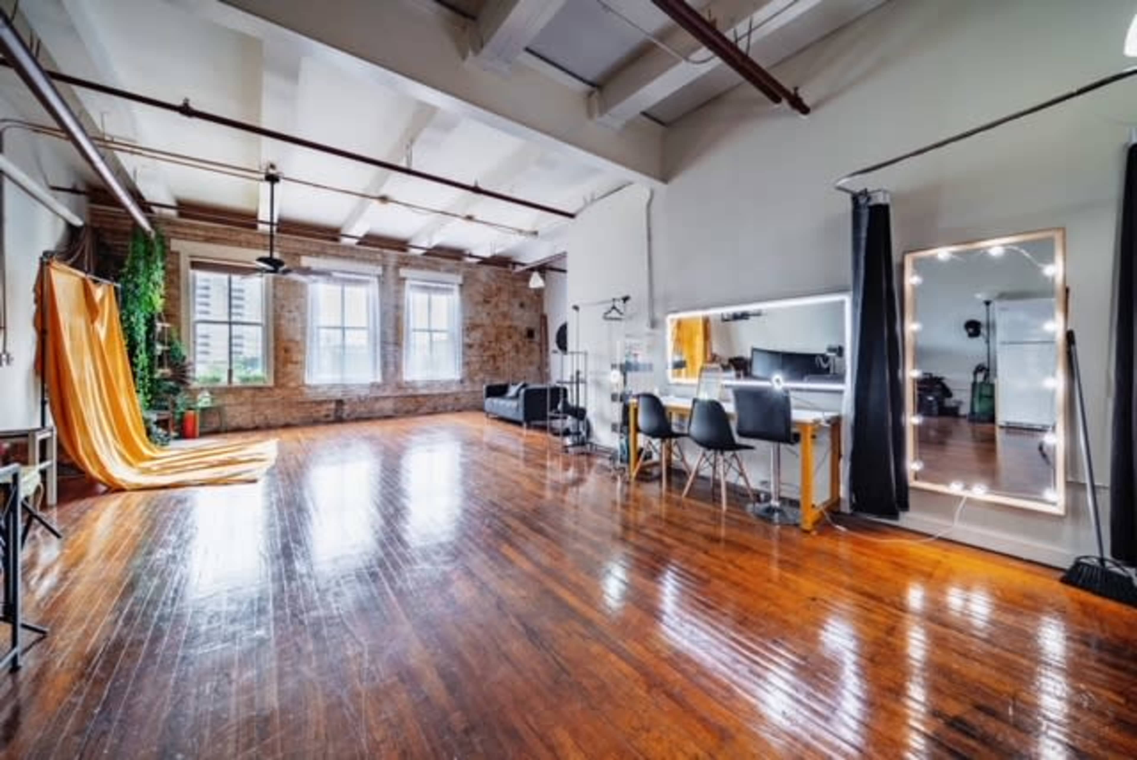 A spacious room with wooden floors, large windows, and areas for sitting, grooming, and creative activities, highlighted by an orange backdrop and a mirror with lights.