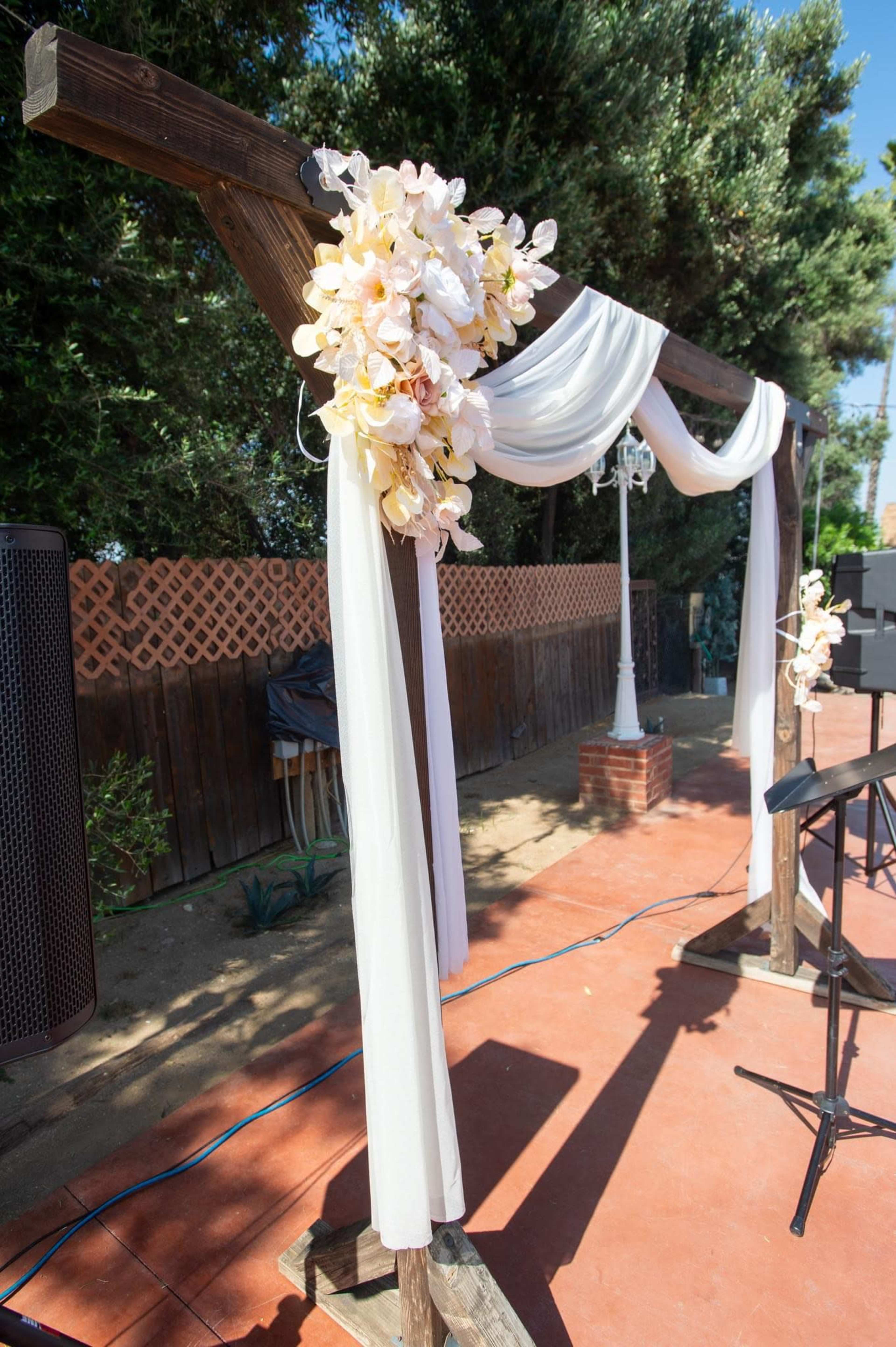 The image shows a wedding arch adorned with white drapes and floral arrangements, set in an outdoor venue.