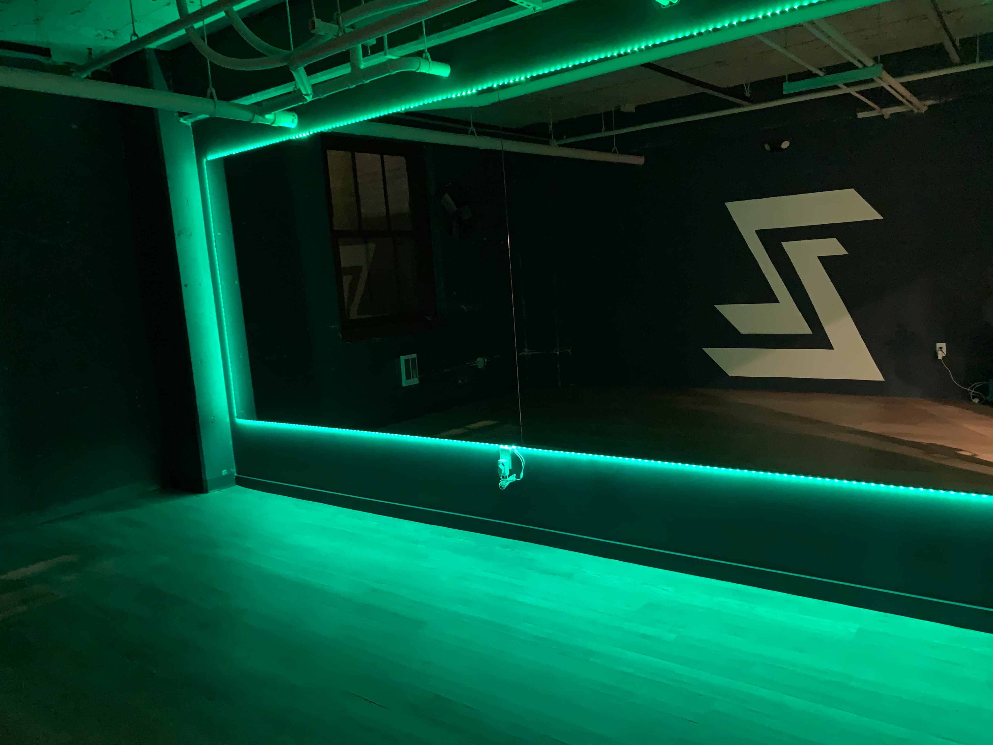 The image shows a dimly lit room with a large illuminated mirror framed in green LED lights and a geometric design on the dark wall.