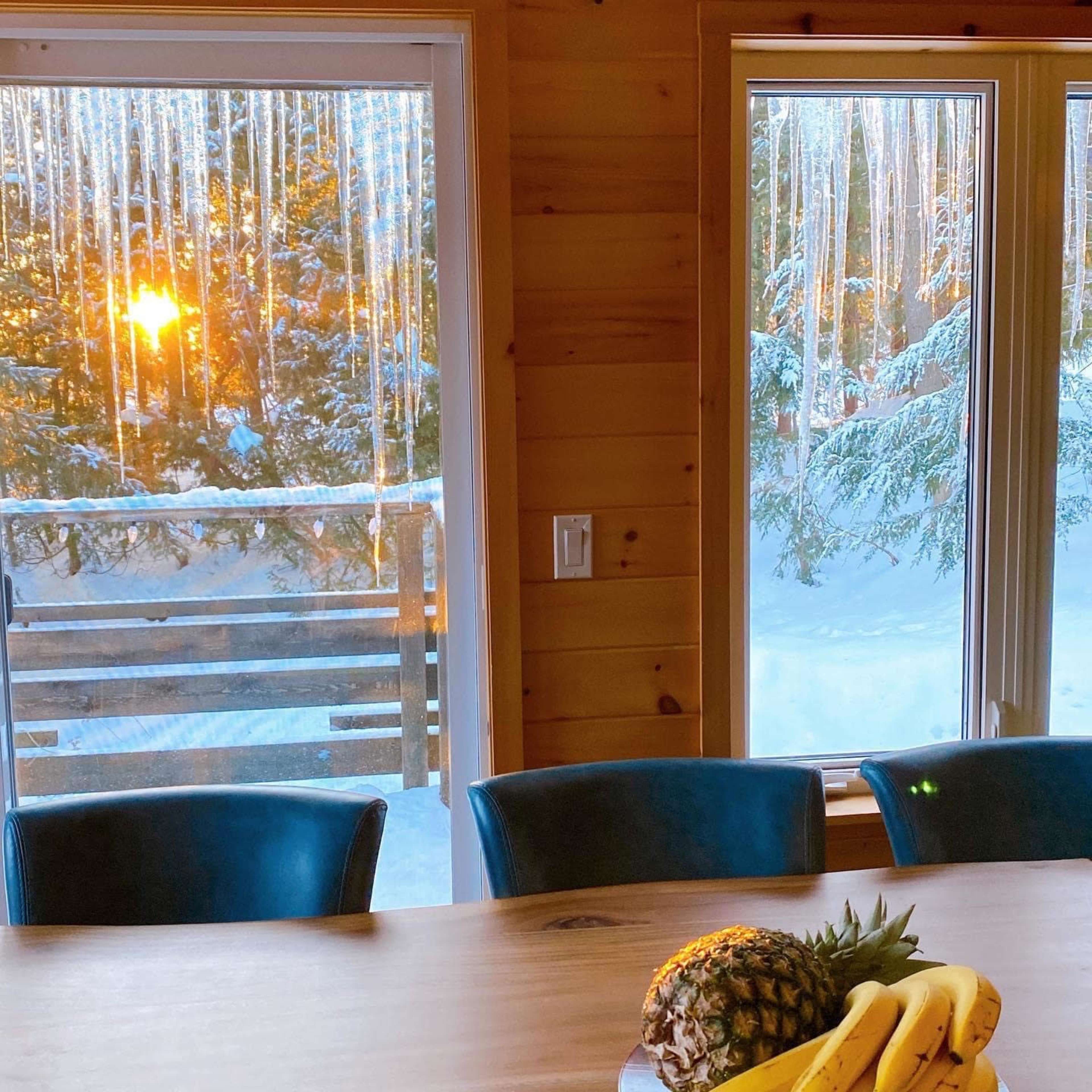 The scene features a wooden dining table with a pineapple and bananas, framed by windows showing icicles and a winter landscape at sunset.