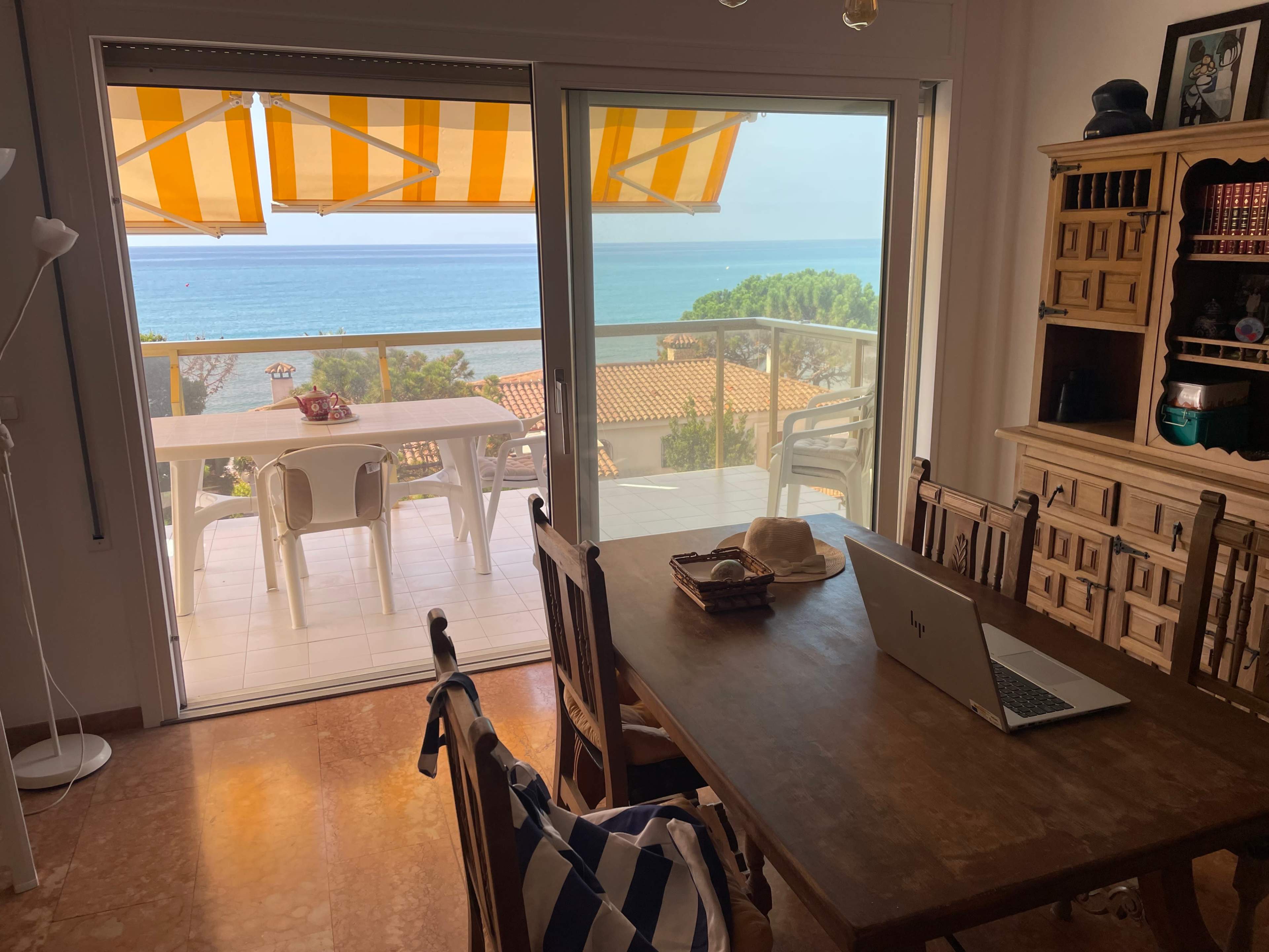 A dining area features a wooden table with chairs, an open door leading to a balcony overlooking the ocean, and a laptop placed on the table.