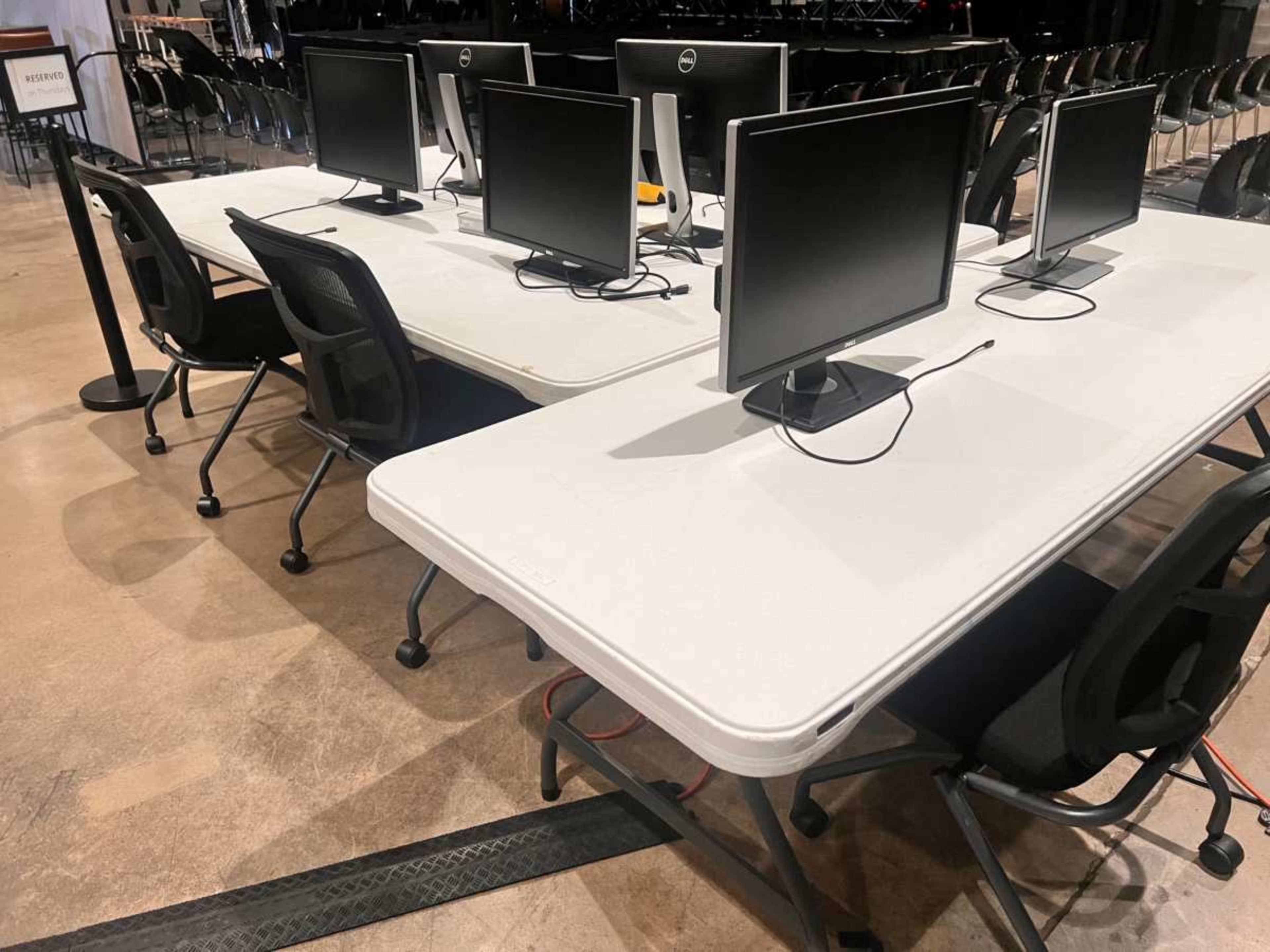 A set of four computers with monitors is arranged on two folding tables in a spacious room with rows of chairs in the background.