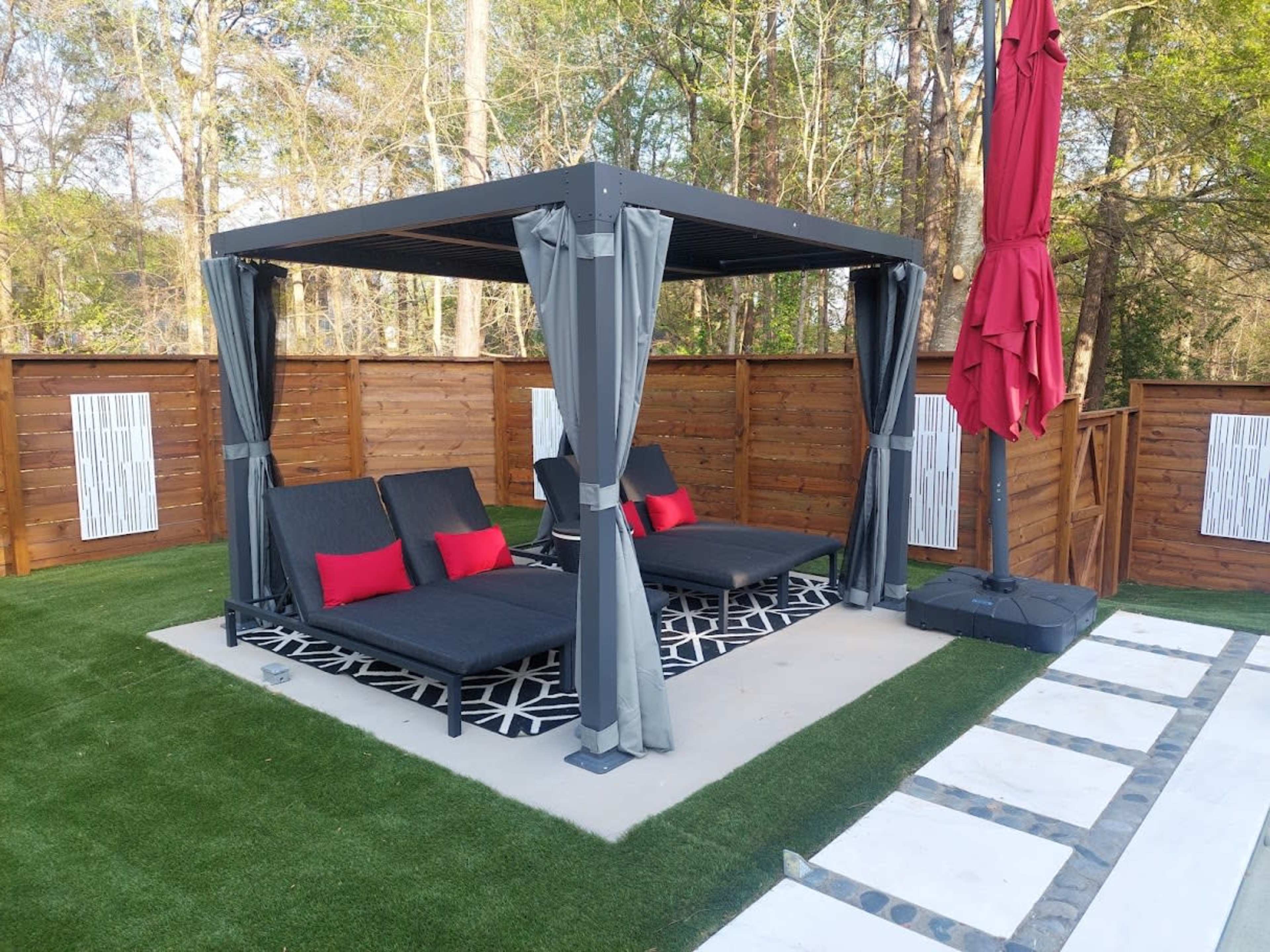 The image shows a shaded outdoor lounging area with two black lounge chairs, red cushions, surrounded by a grassy lawn and wooden privacy fencing.