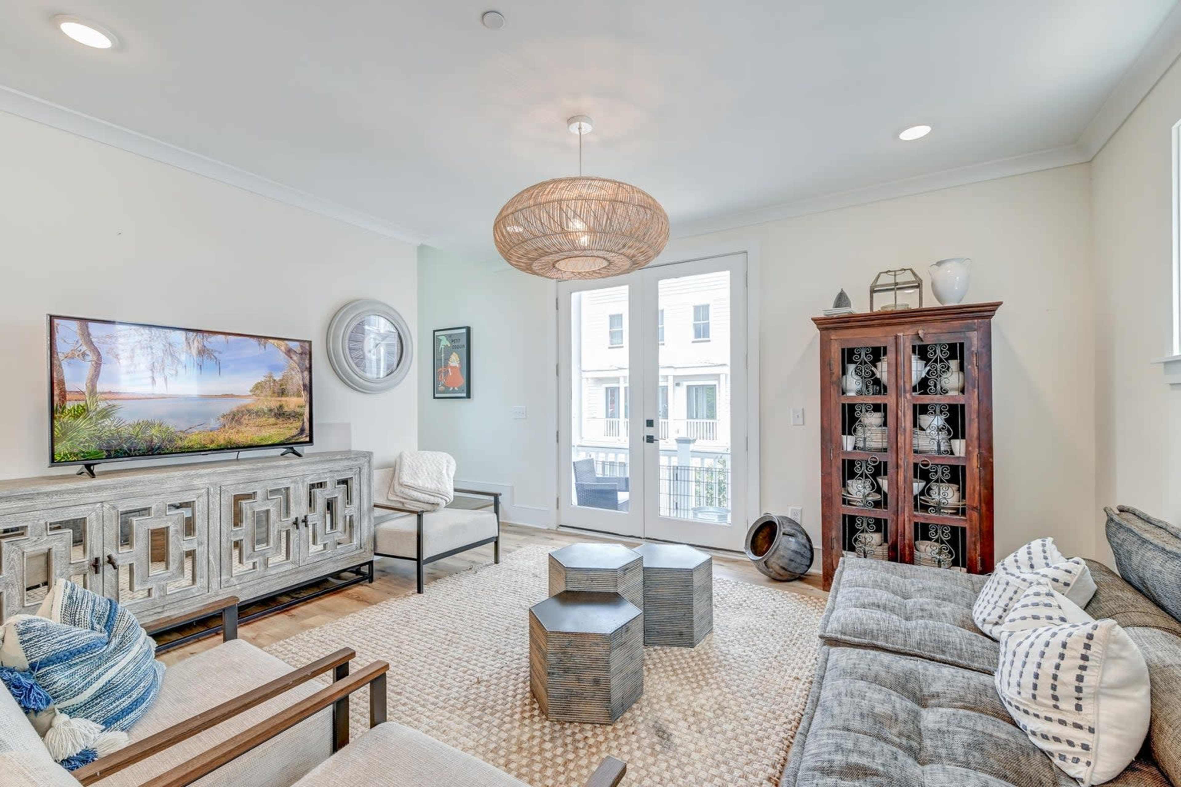 A modern living room with a television, a set of wooden furniture, a woven light fixture, and large windows leading to a balcony.
