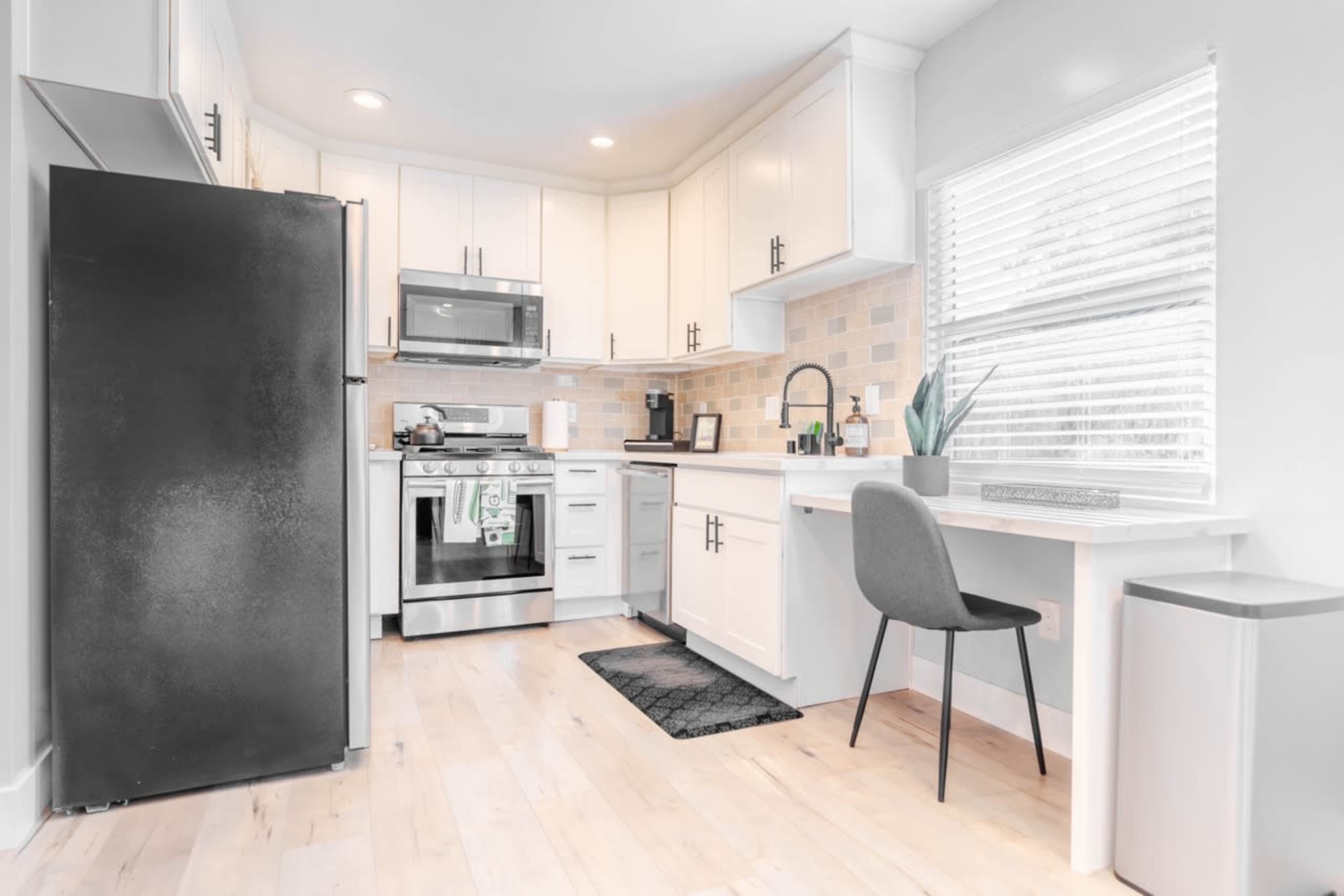A modern kitchen features stainless steel appliances, white cabinetry, and a small dining area with a desk by the window.